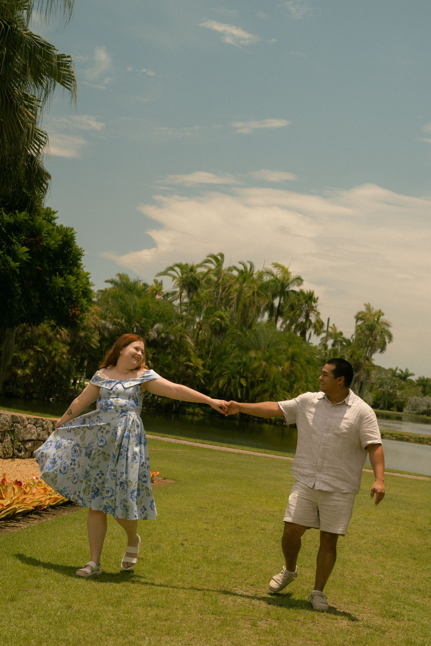 A couple holds hands and dances together on a green lawn surrounded by tropical palm trees under a bright blue sky.