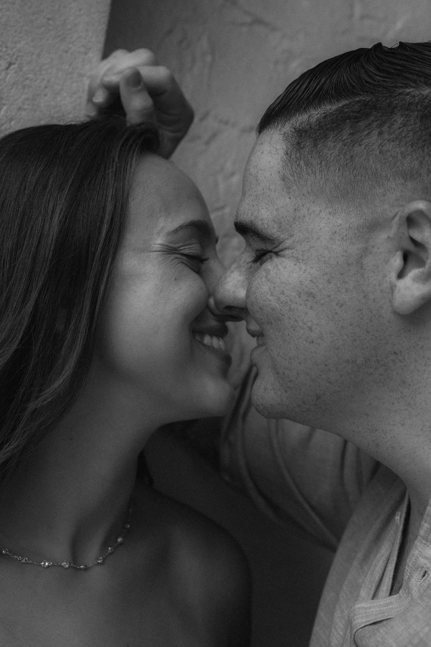 Black and white photo of a couple sharing a romantic kiss, smiling closely together against a textured wall.