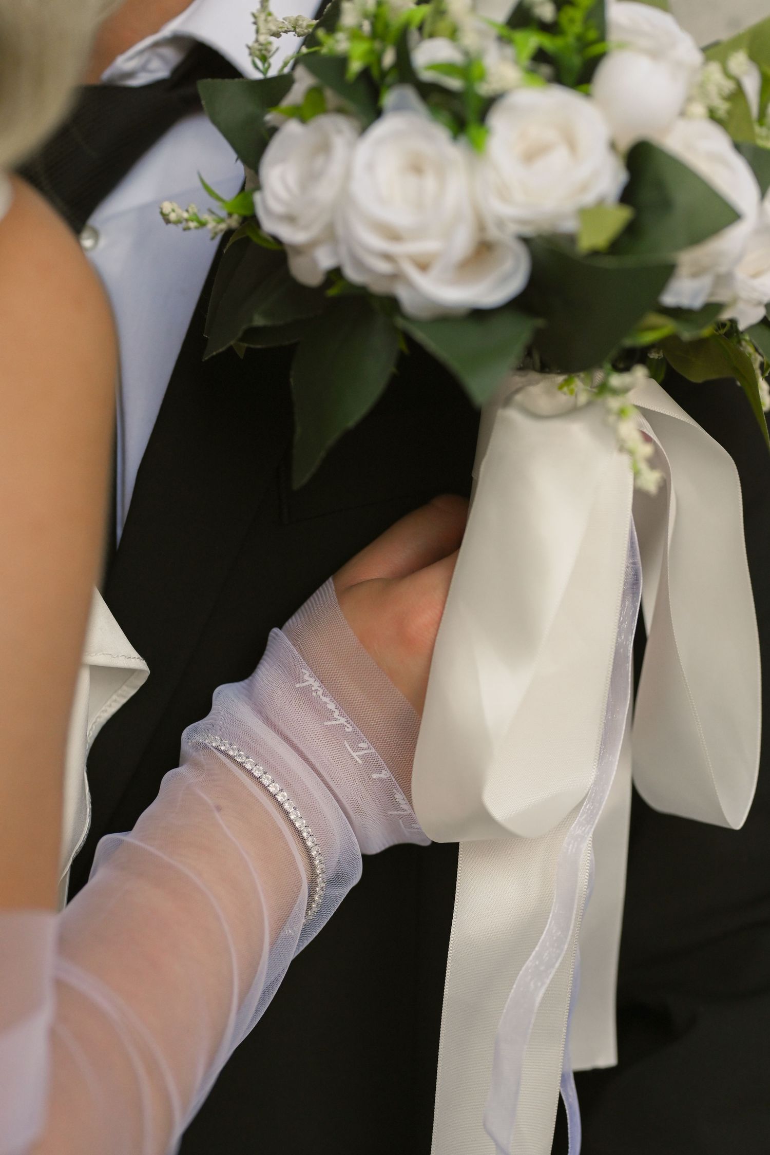 Close-up of a white bridal bouquet with roses and ribbons being held by gloved hands.