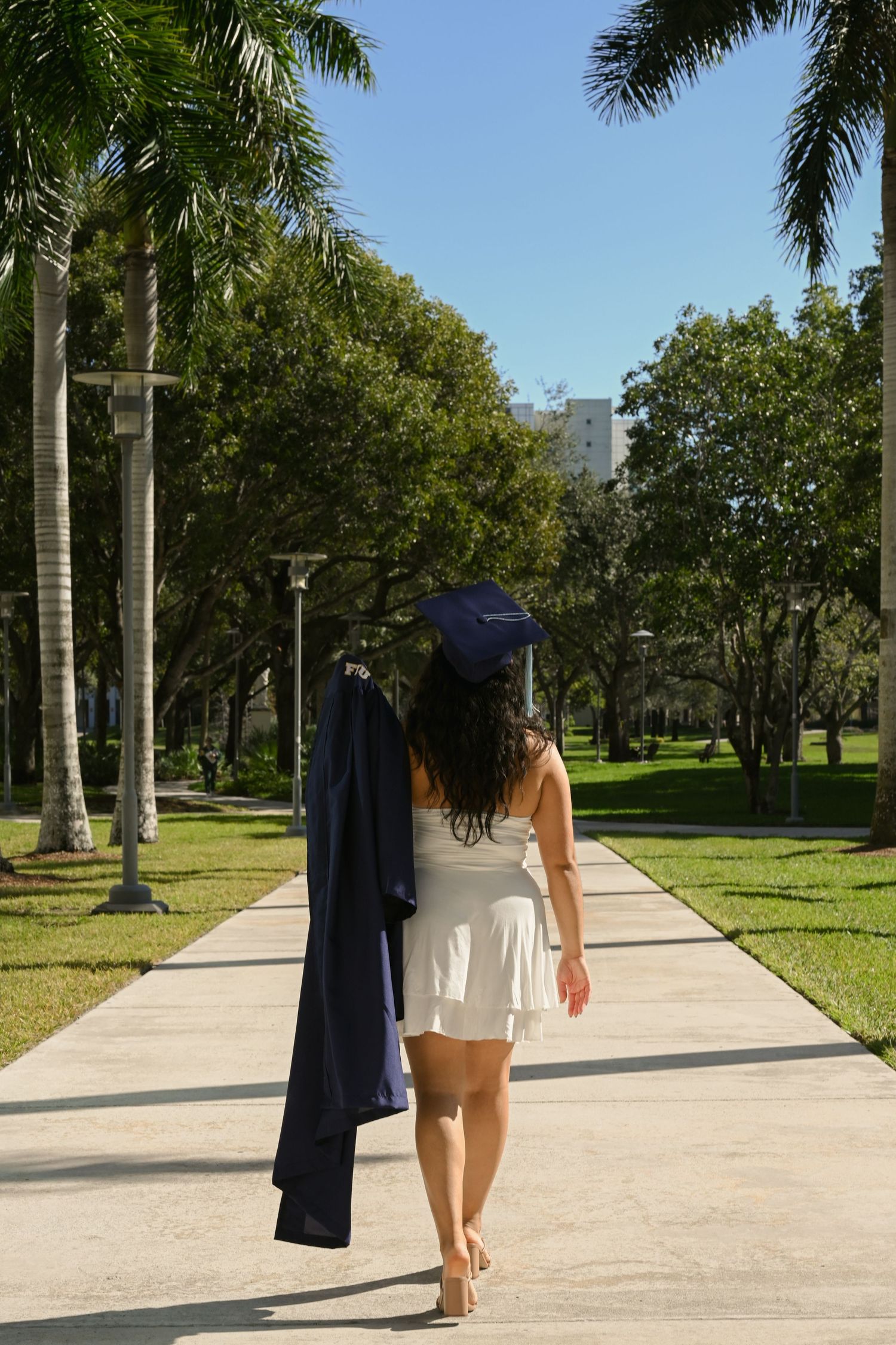 A graduate in navy blue gown walks along a palm tree-lined pathway on a sunny day.