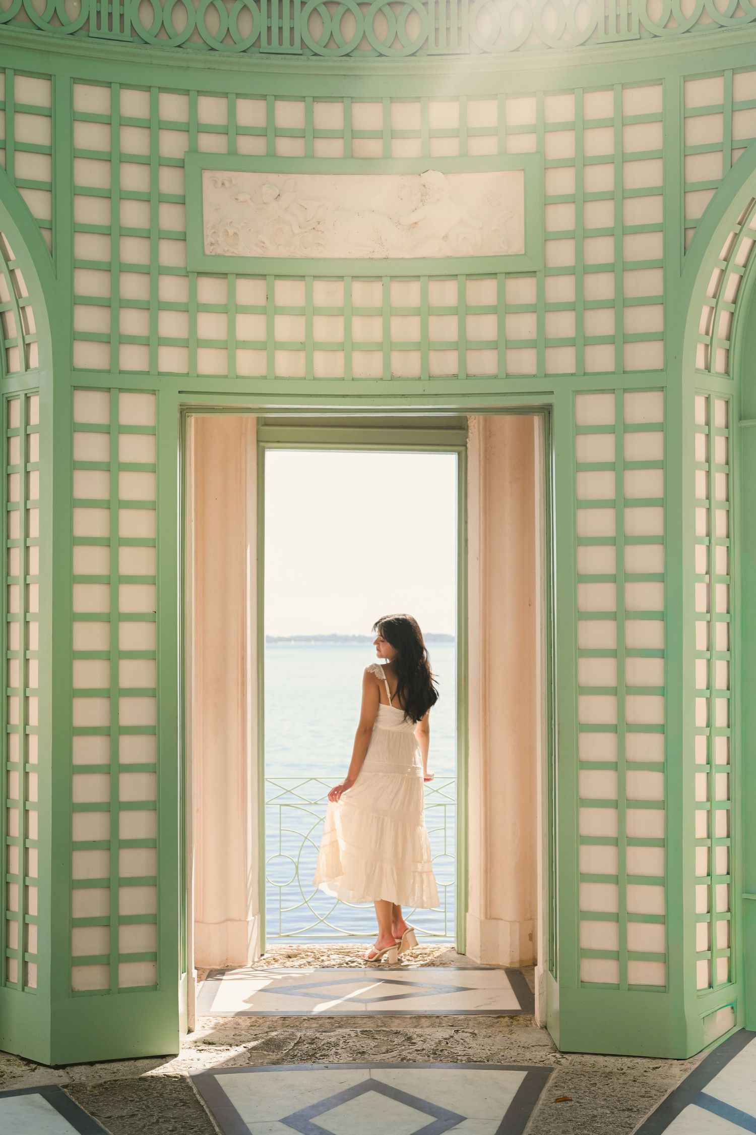 Woman in white dress standing in elegant green and white archway overlooking serene ocean view.