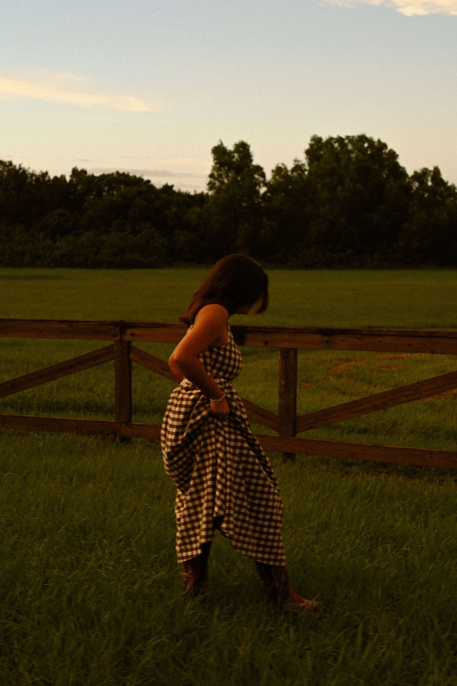 A black and white gingham dress flows in the breeze against a rustic wooden fence at sunset.