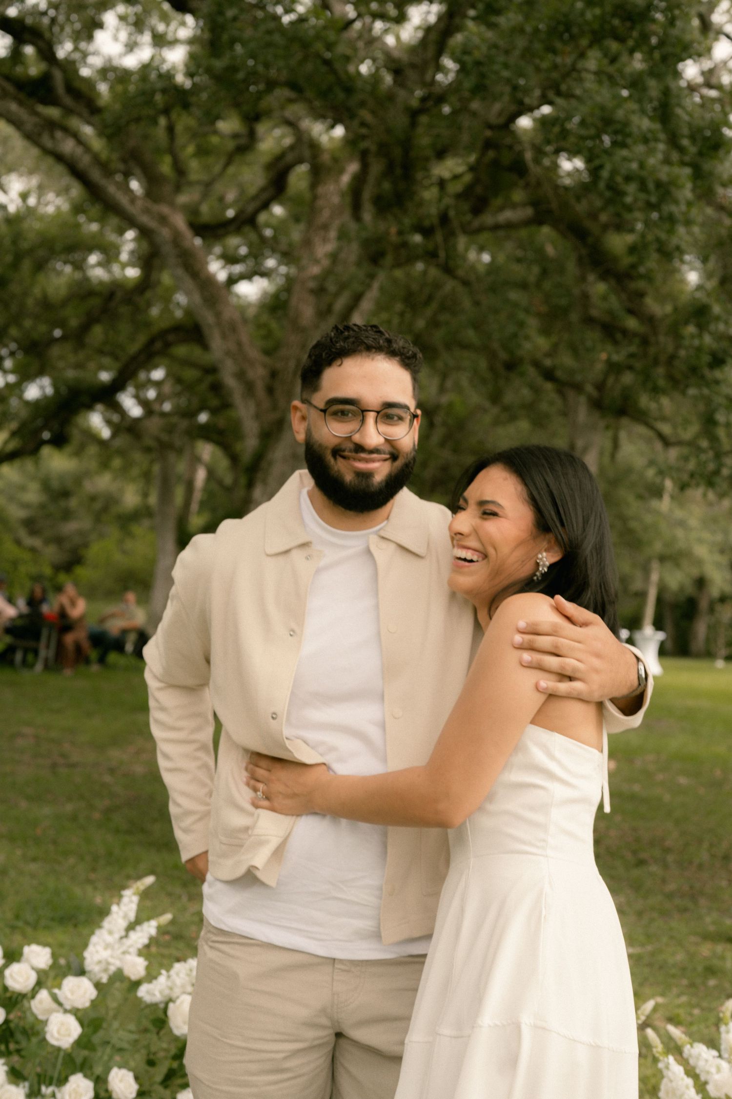 Happy couple embracing outdoors under large oak trees, man in beige jacket and woman in white dress.