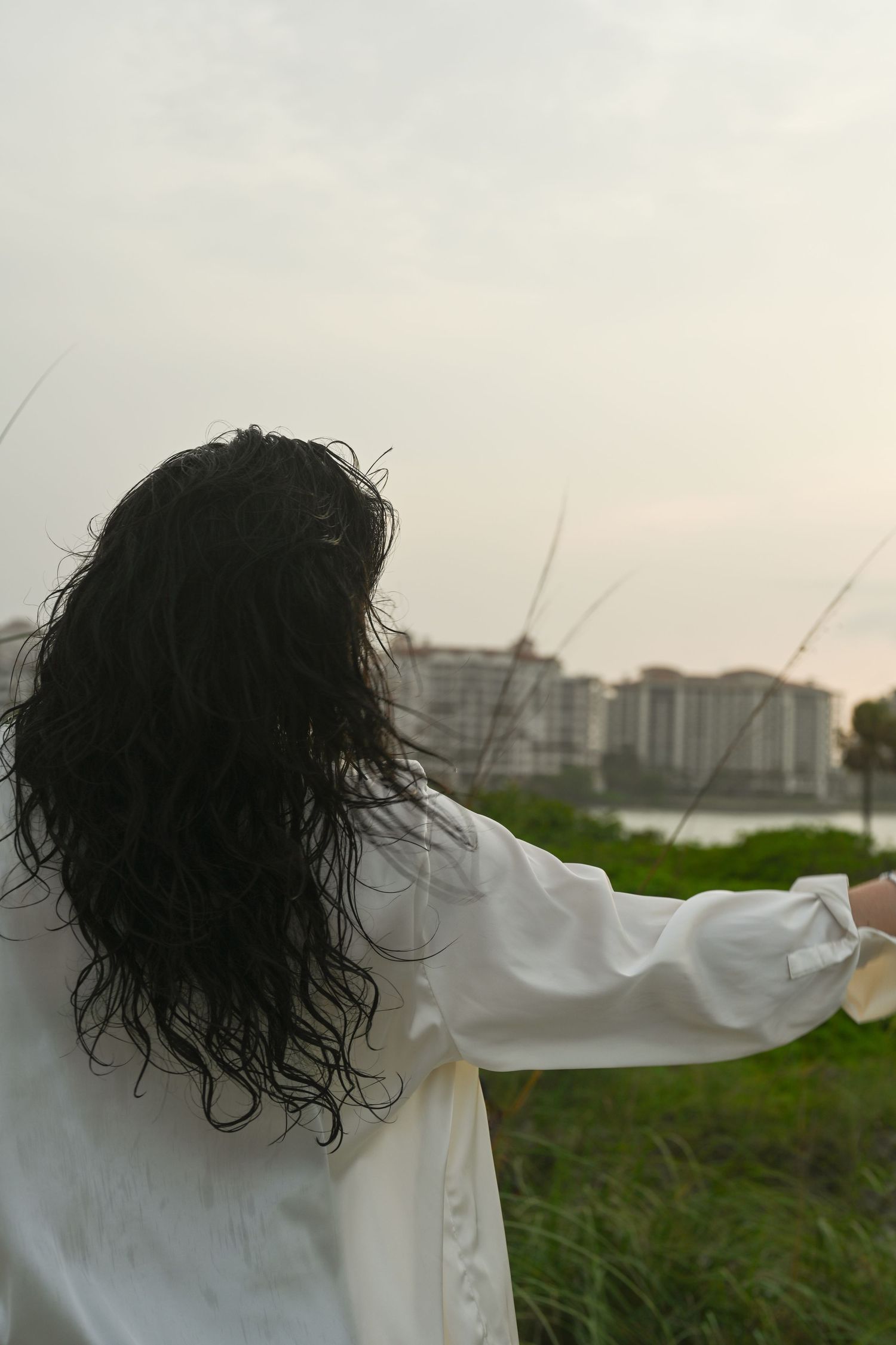 Figure in flowing white shirt poses in grassy field with cityscape background.