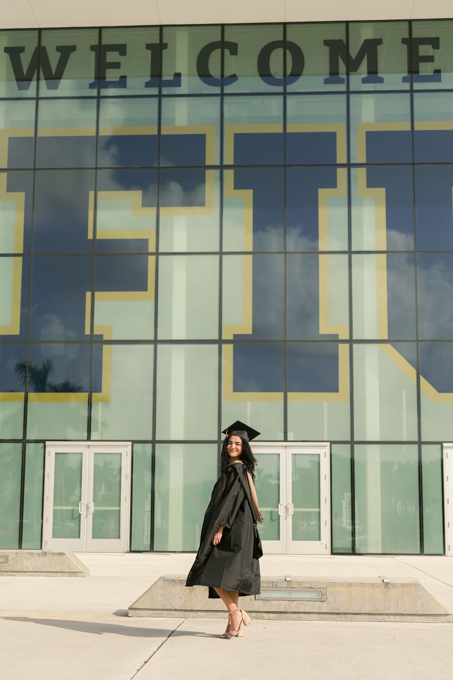 Graduate stands in front of glass building with 'WELCOME' signage.