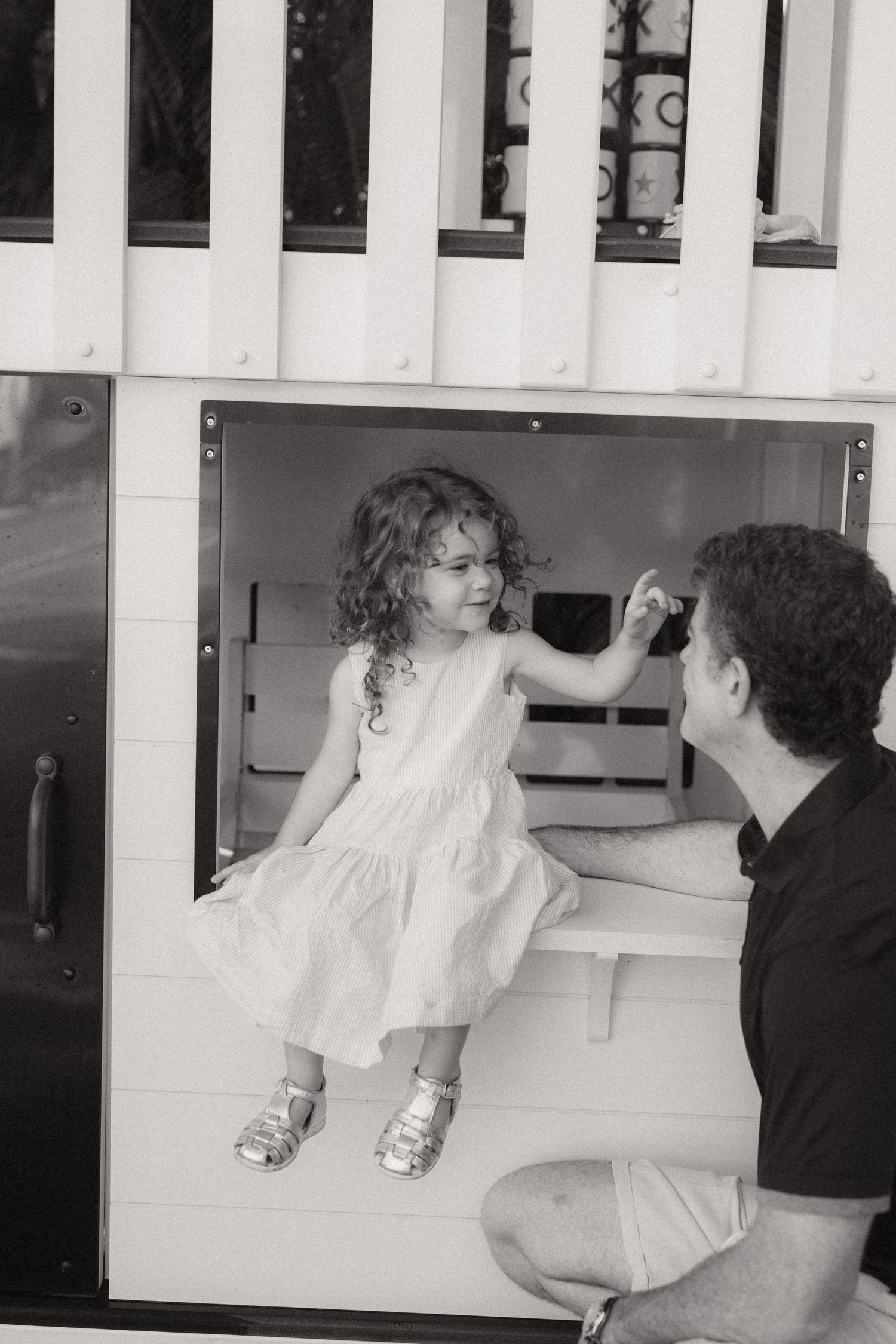 Black and white photo of a curly-haired little girl in a dress sitting in a playhouse window, reaching toward a smiling man.