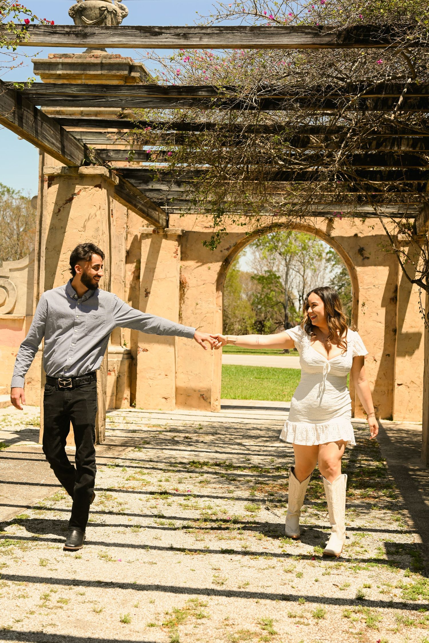 Playful couple holds hands while dancing under rustic pergola structure.