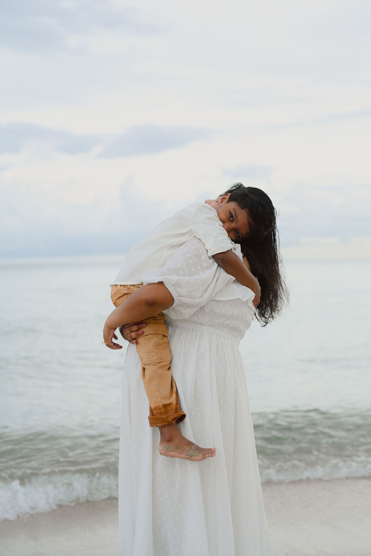 A tender moment captured on a beach as a figure in a flowing white dress holds someone in khaki pants against the ocean backdrop.