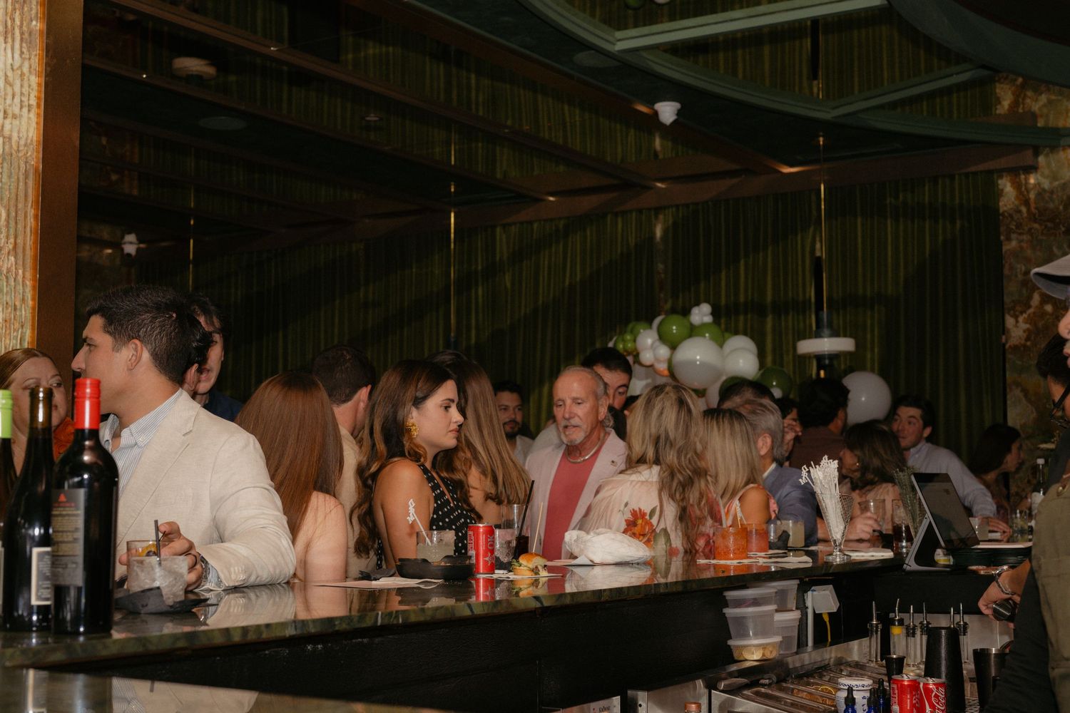 Guests socializing at a dimly lit bar with green drapes, wine bottles, and white balloon decorations in the background.