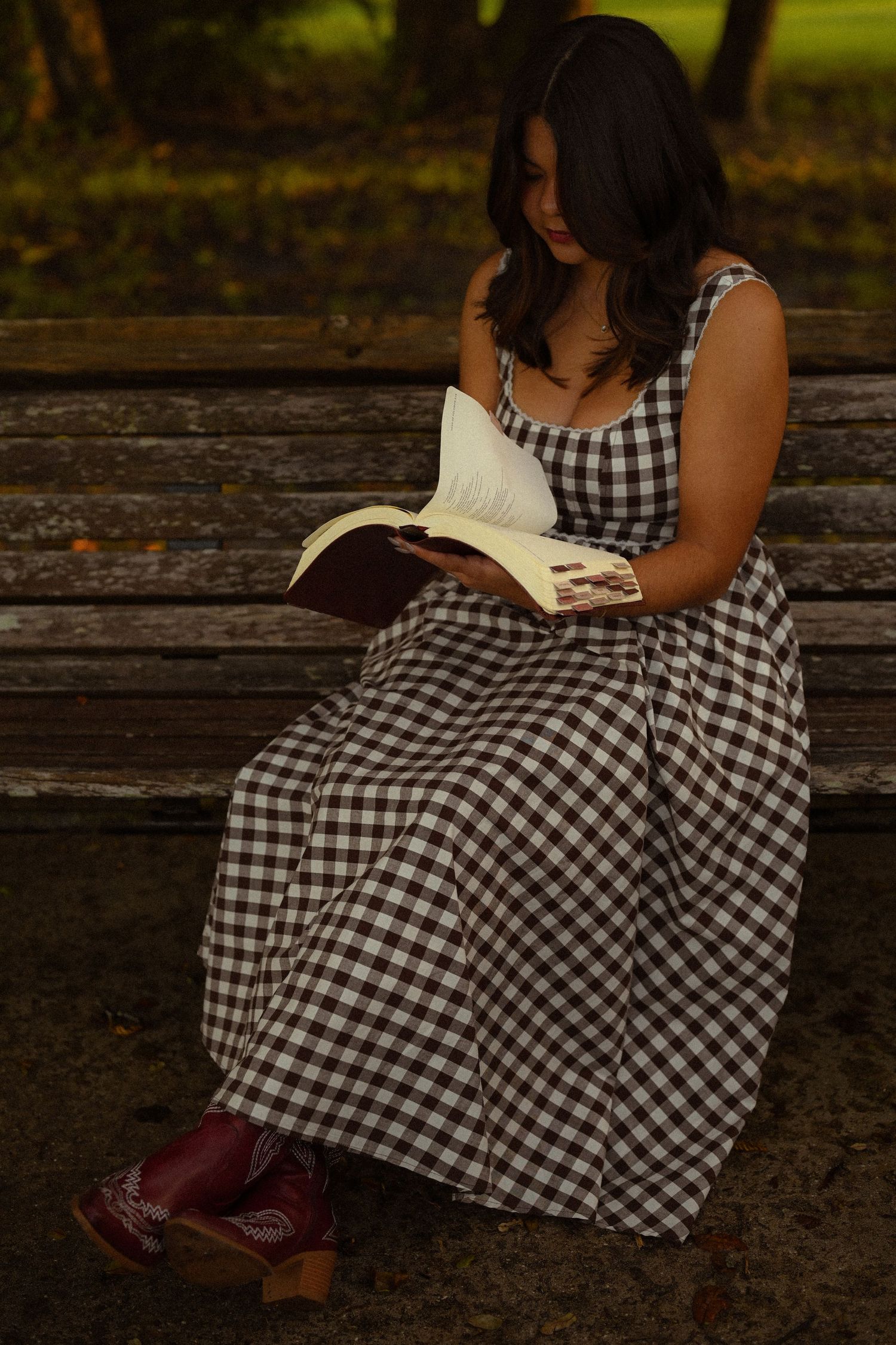 Someone wearing a black and white checkered dress sits on a rustic wooden bench with a book.