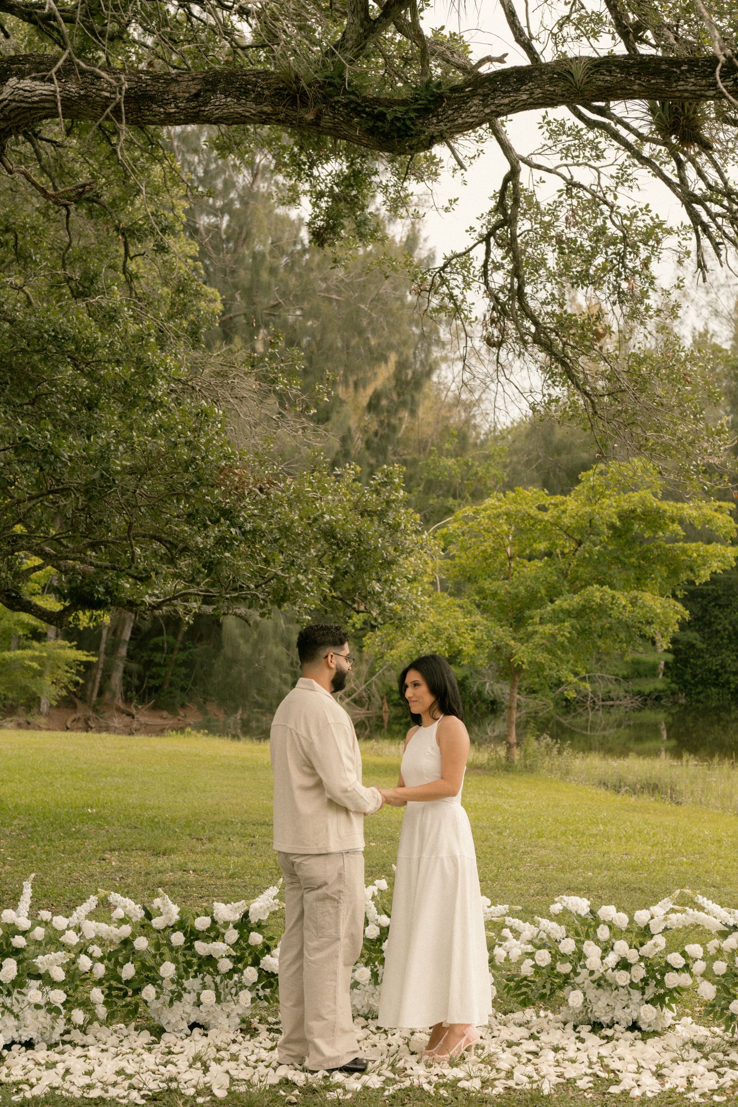 Couple holding hands under oak tree surrounded by white flowers during outdoor wedding ceremony.