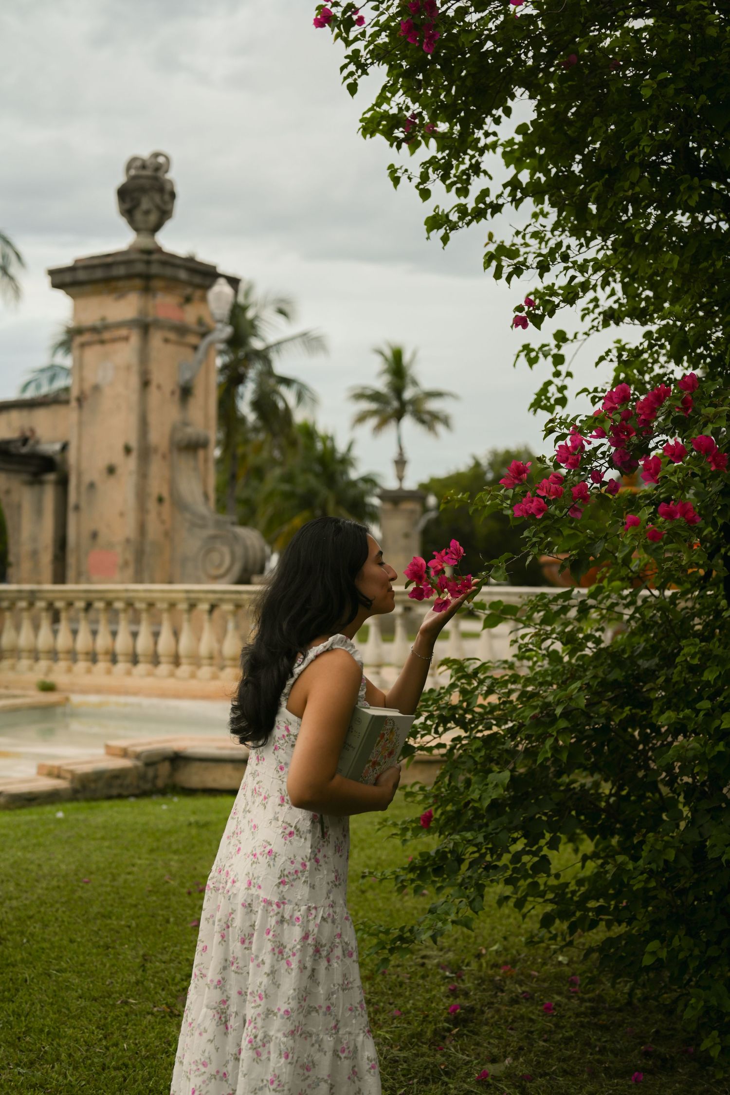 A person in a white floral dress poses near pink bougainvillea flowers and stone architecture in a garden setting.