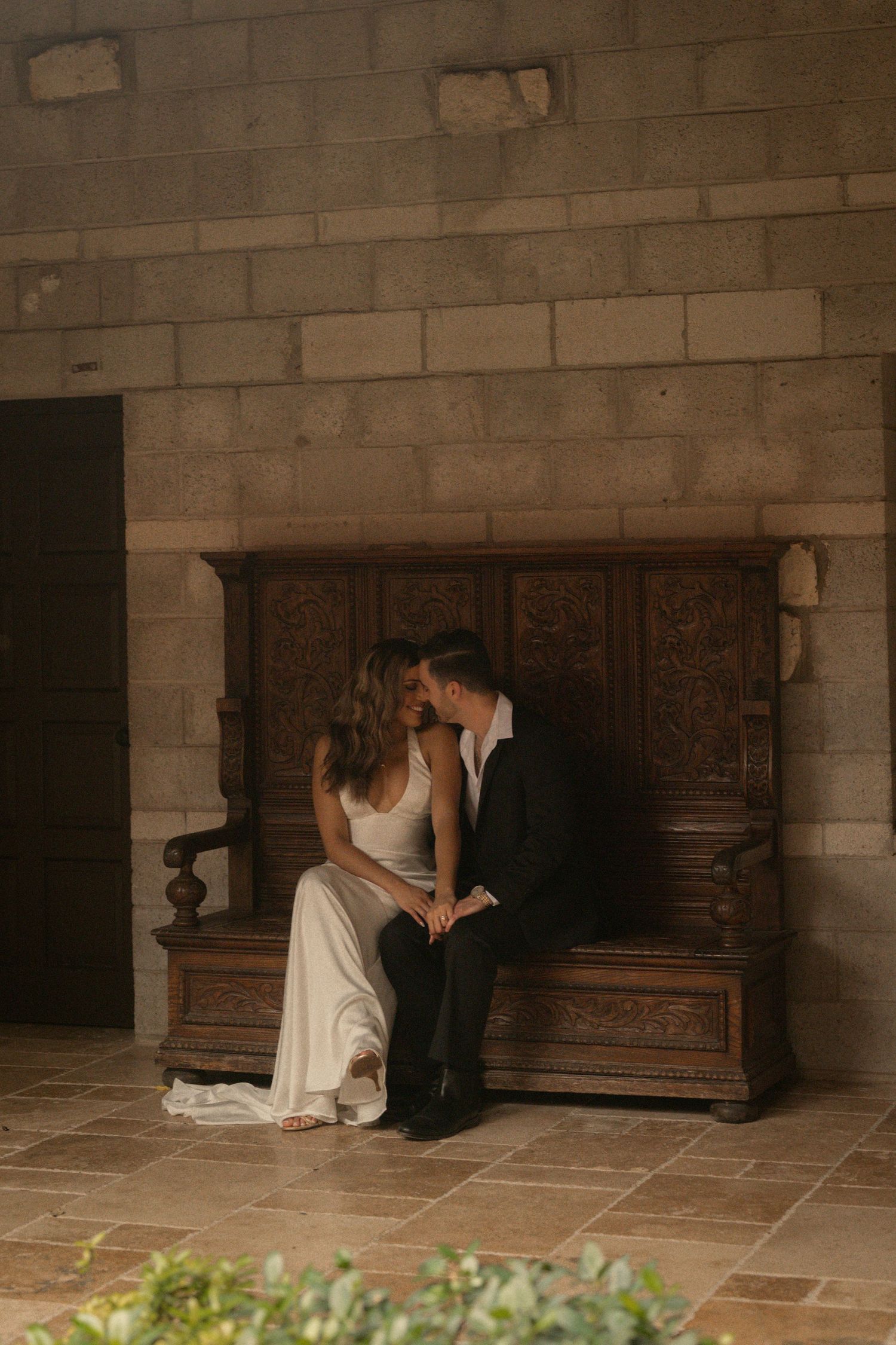 Couple in a white gown and dark suit sit close together on an ornate carved wooden bench against a rustic stone wall.