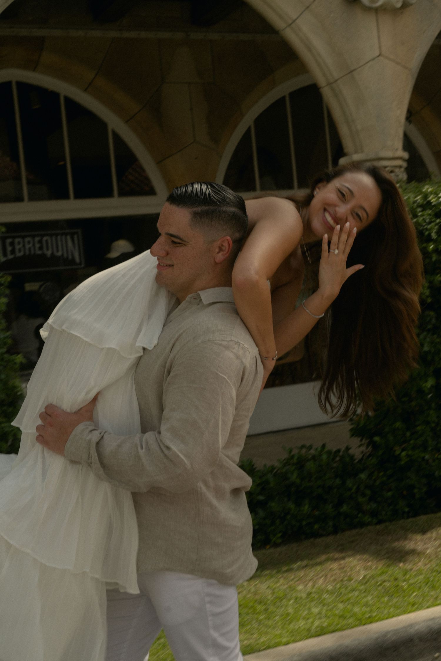 A smiling couple in white outfits pose playfully outdoors near arched windows, with the woman leaning on the mans back.