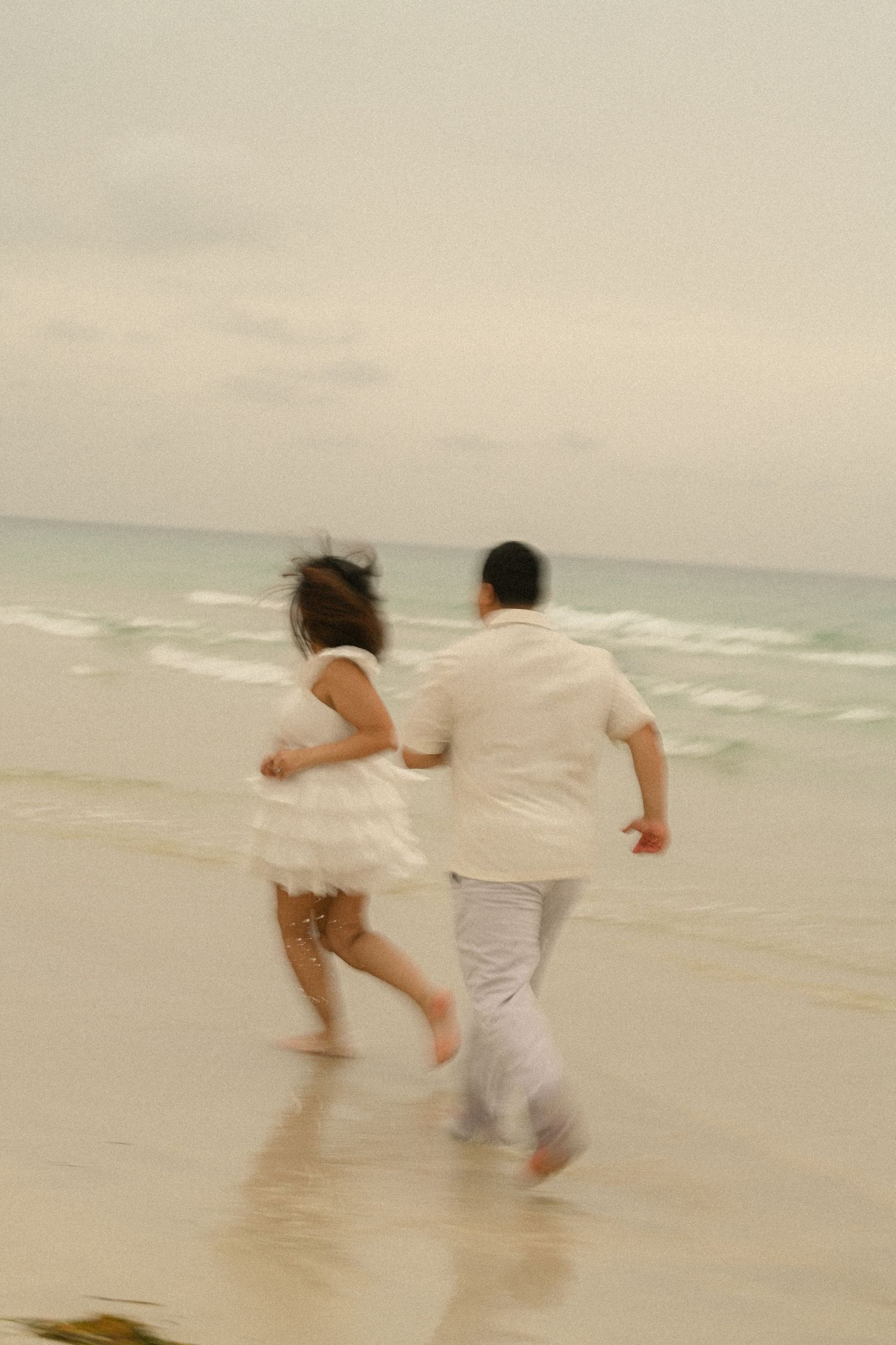 A couple in white clothing runs playfully along the shoreline with waves crashing behind them.