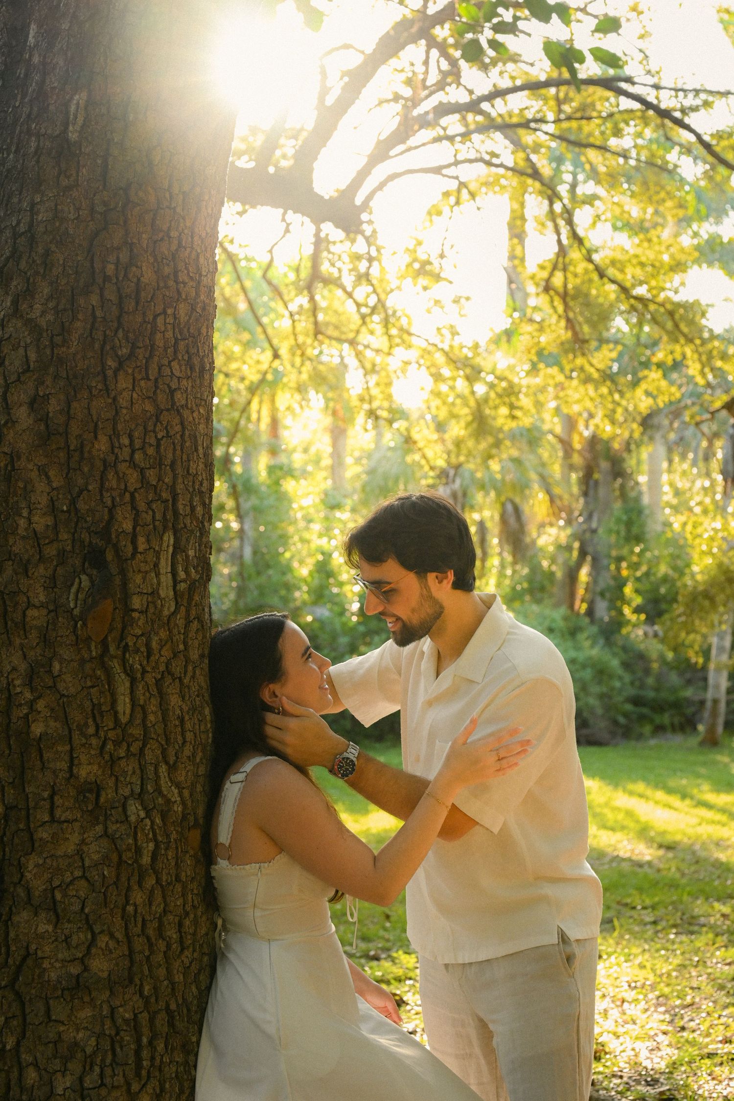 A couple in white outfits shares a playful moment in a sunlit forest setting during golden hour.