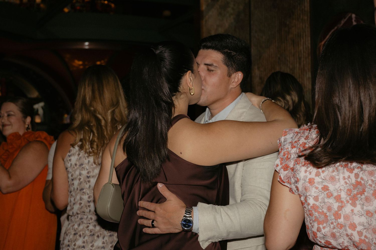Couple sharing a romantic embrace and kiss at a lively indoor social event or wedding reception.