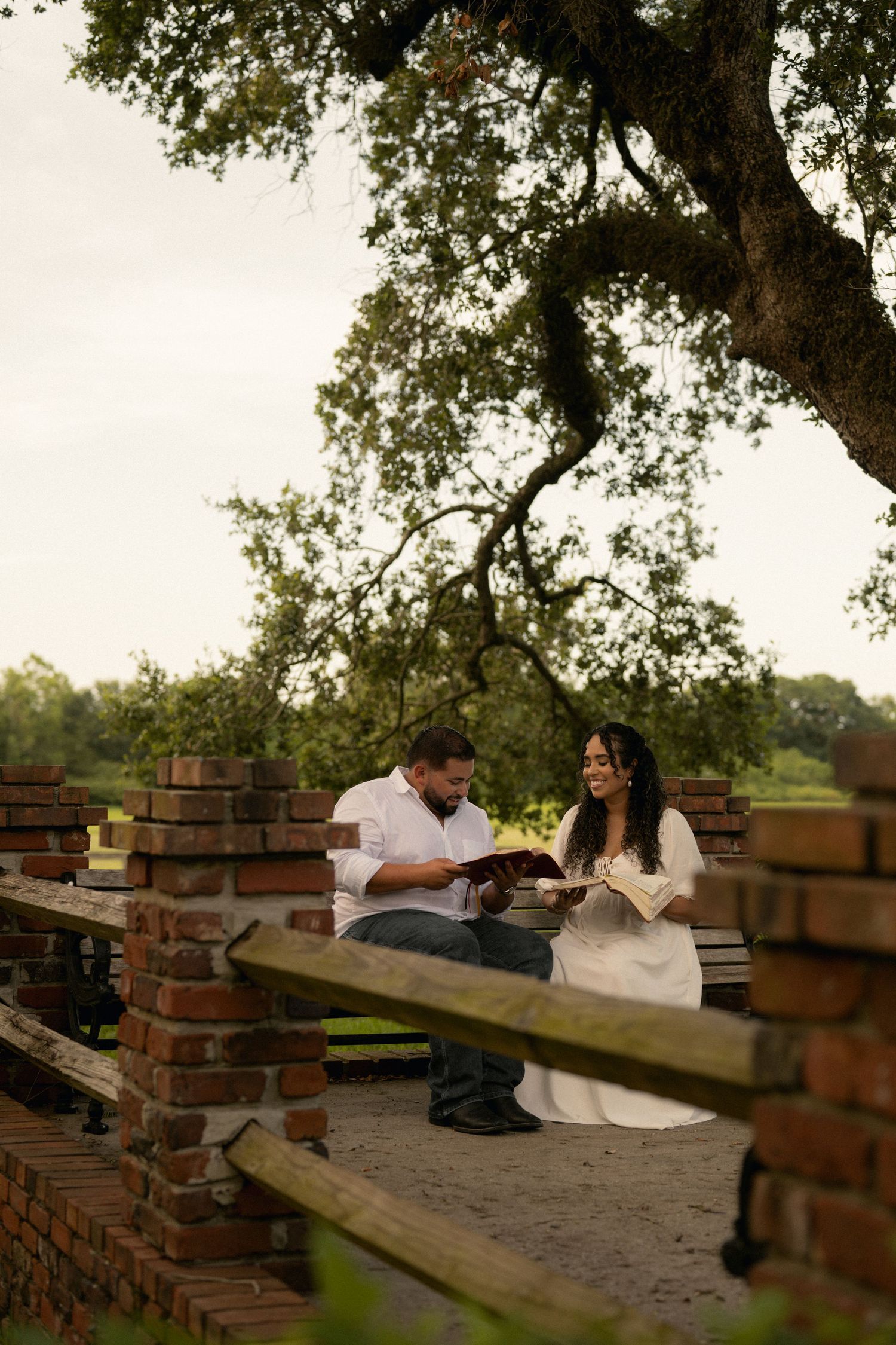 Couple sitting on a bench under a large oak tree, sharing books and smiling in an outdoor brick setting.