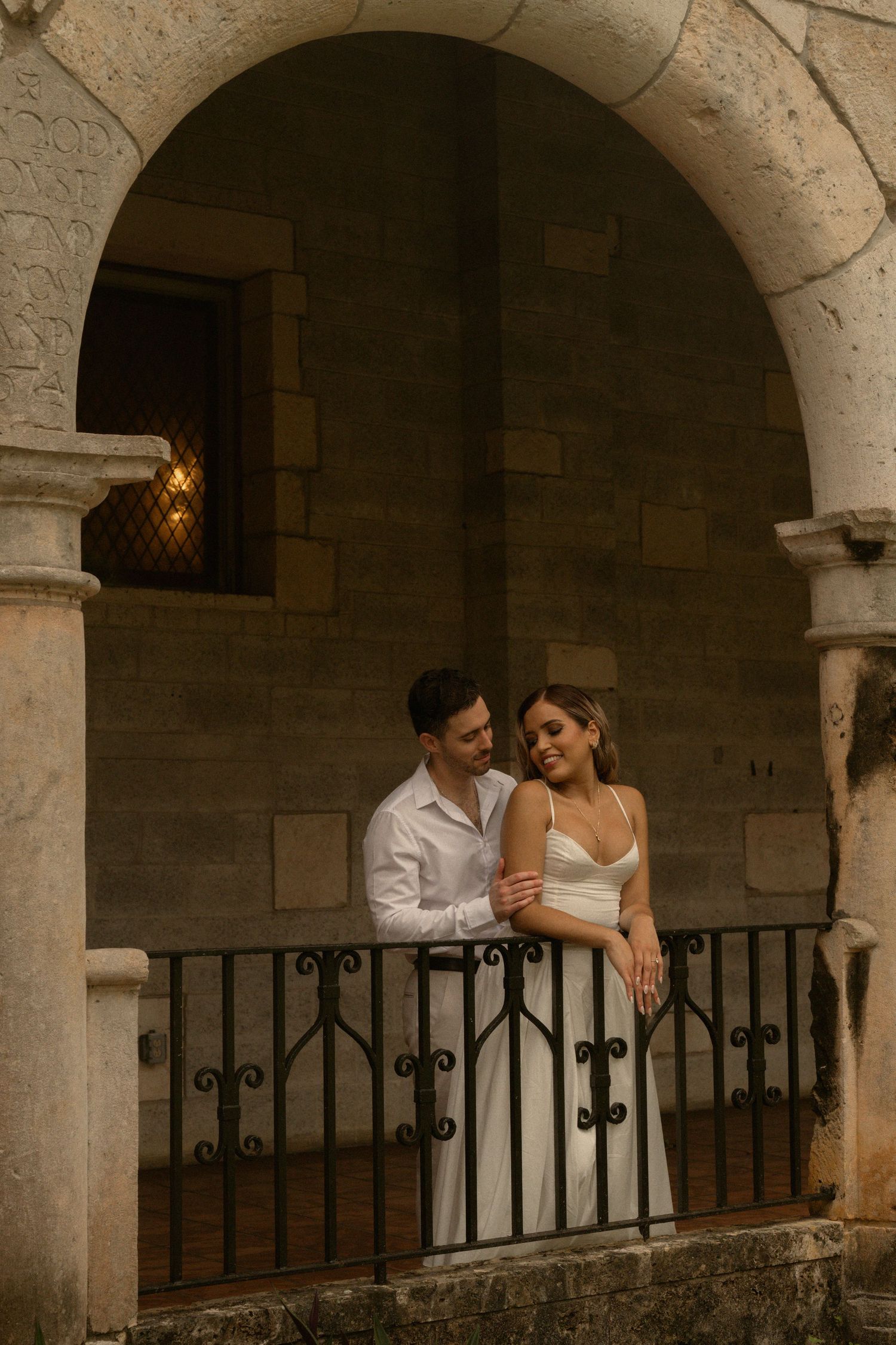 Couple in white embracing on an ornate wrought iron balcony beneath a rustic stone archway during an engagement photoshoot.