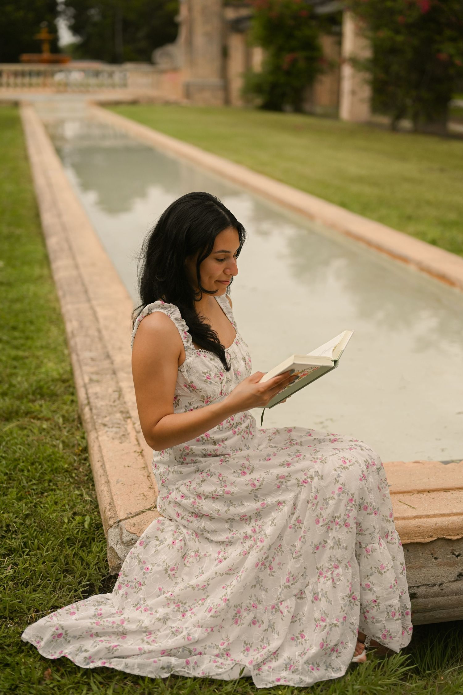 Person in floral dress reads book while sitting on concrete path near manicured lawn.