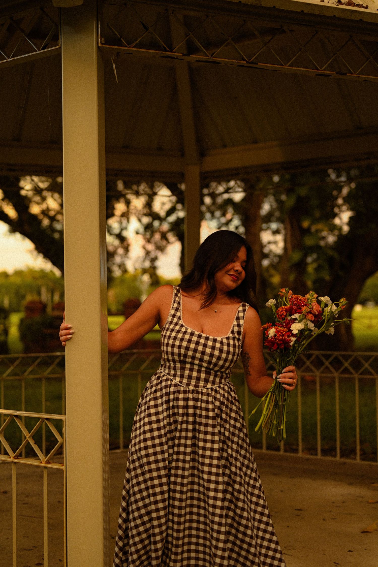 Person wearing checkered dress holds flowers while posing inside an outdoor pavilion.