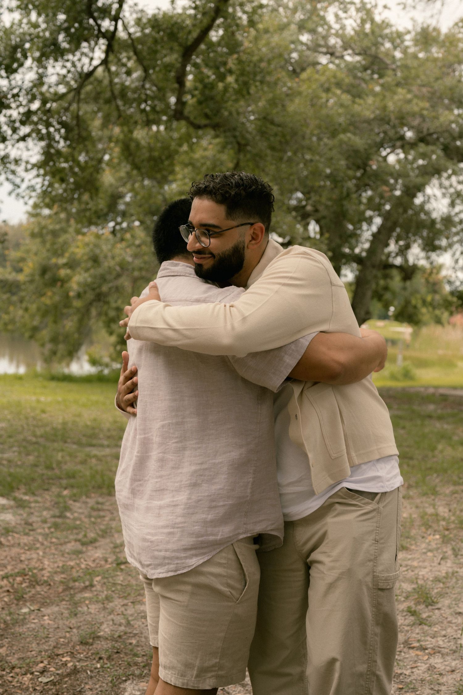 Two men share a warm embrace outdoors near a lake surrounded by green trees.