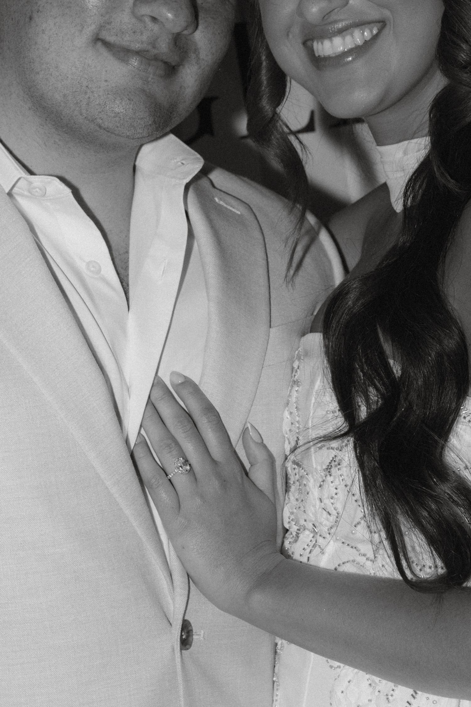 Black and white photo of a couple smiling, woman showing off engagement ring on her hand resting on mans chest.
