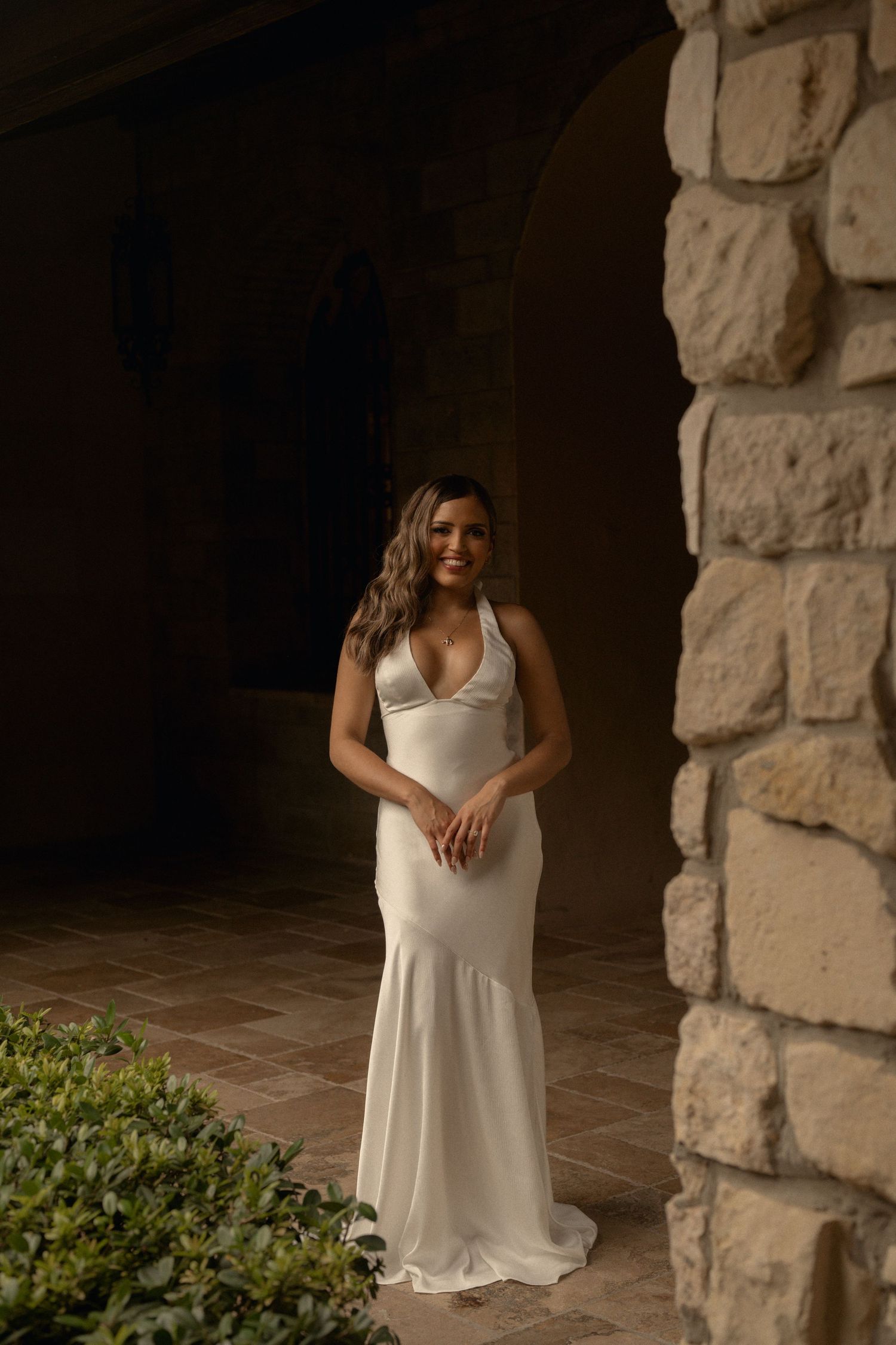 Smiling bride in an elegant white halter gown stands beside a rustic stone wall in a shaded courtyard with warm brick flooring.