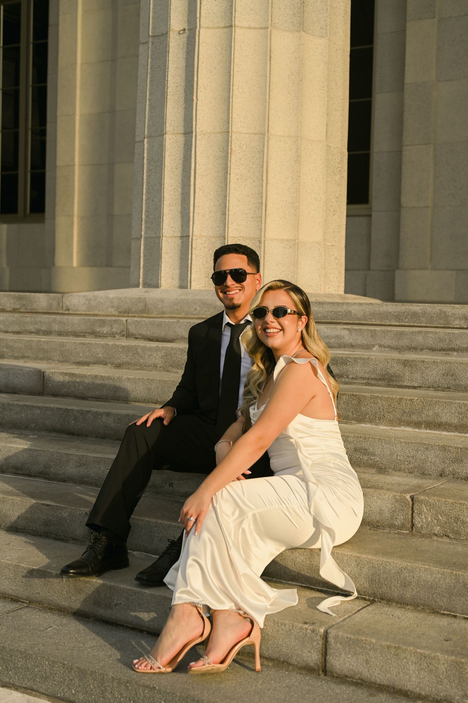Bride in white dress and groom in black suit pose casually on stone steps at sunset.