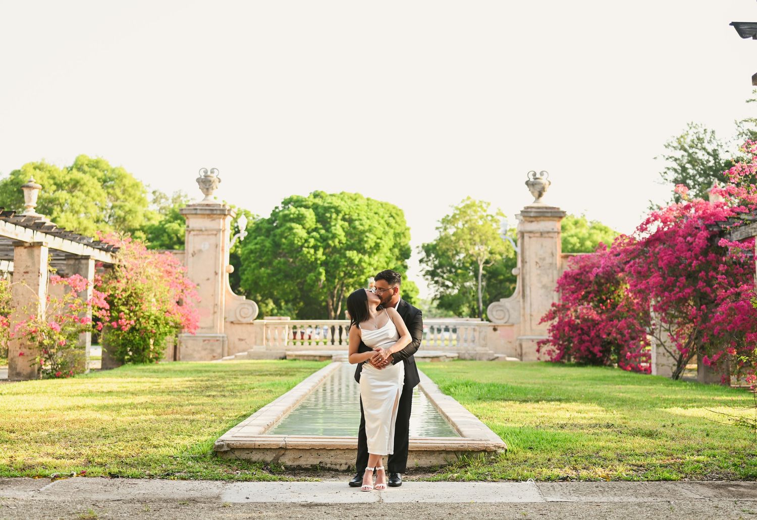 Wide panoramic view of a couple in formal wear standing by stone pillars in a garden setting.