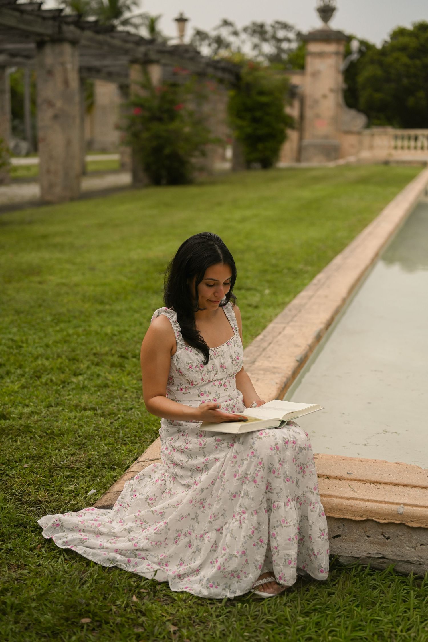 A figure in a white floral dress reads while sitting beside a stone pathway in a garden setting.