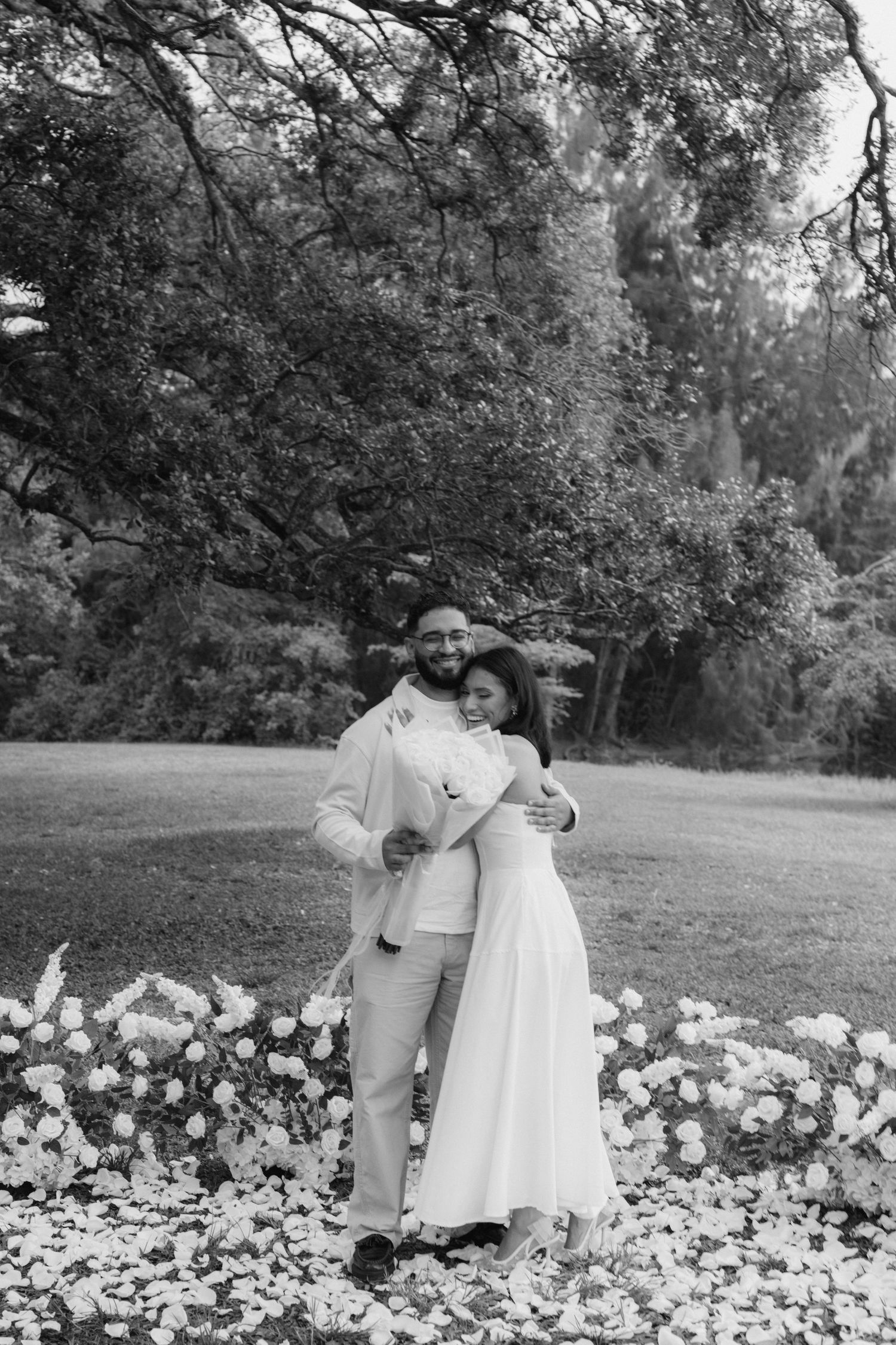 Black and white photo of a couple embracing in a garden, wearing wedding attire, surrounded by white flowers.