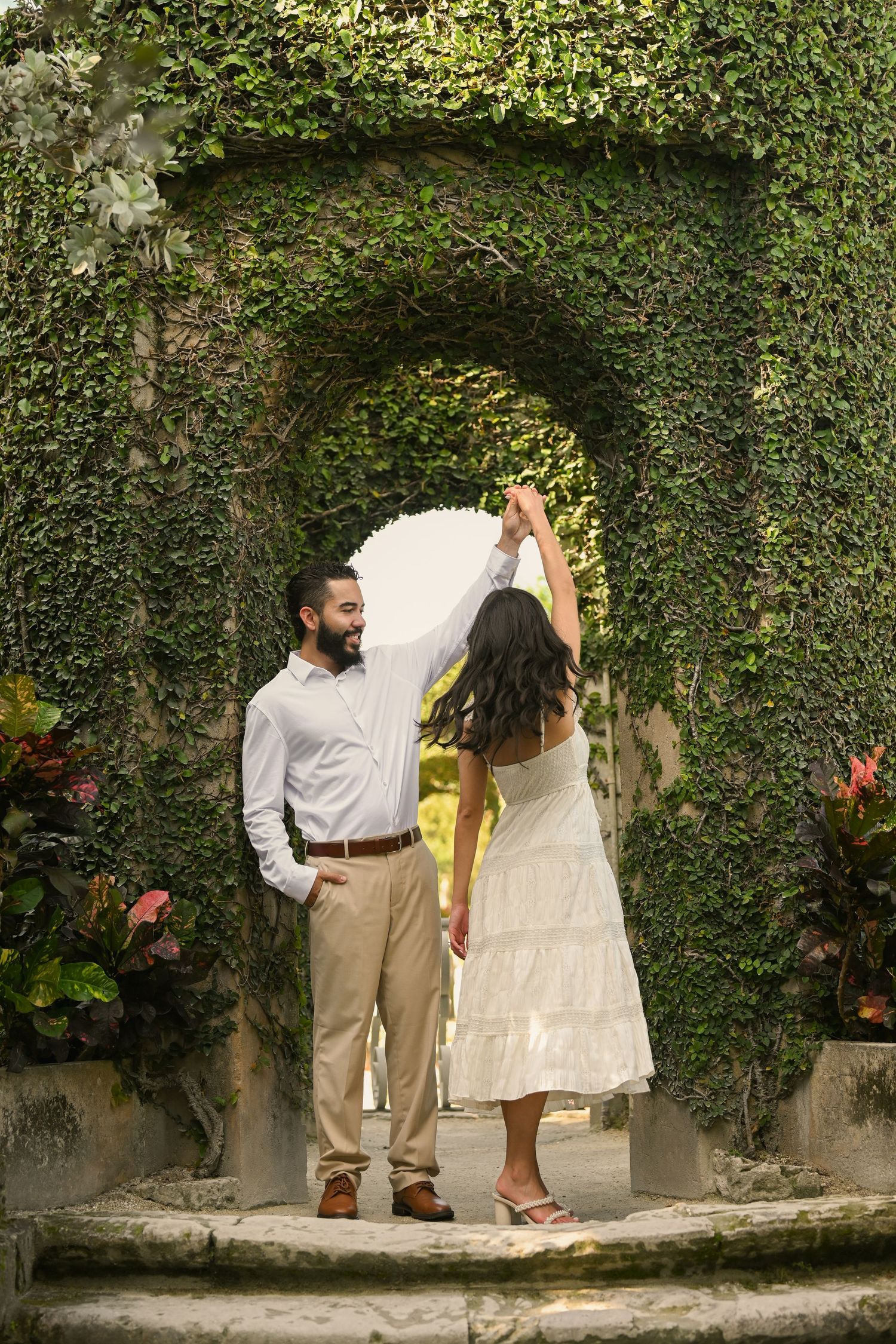 A romantic couple poses together against a lush ivy-covered wall in casual wedding attire.