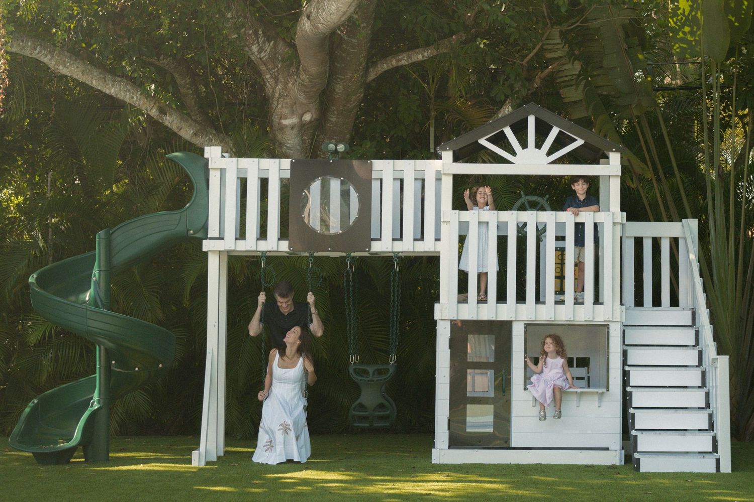 Children play on a white wooden backyard playset with a green spiral slide, swings, and stairs while a woman and man stand beneath it.