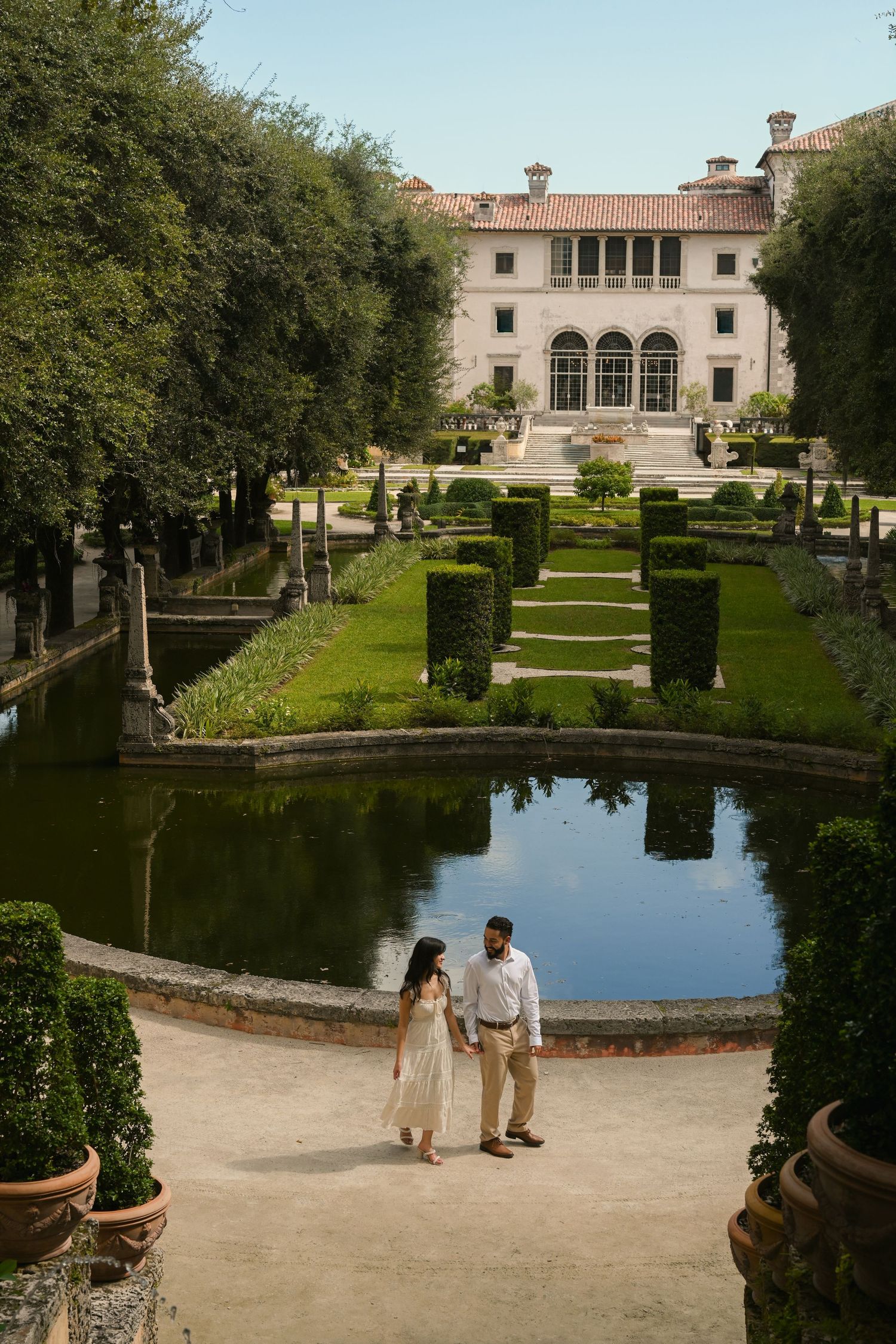 A romantic couple strolls through the manicured gardens of a grand estate with reflecting pools and Italian architecture.