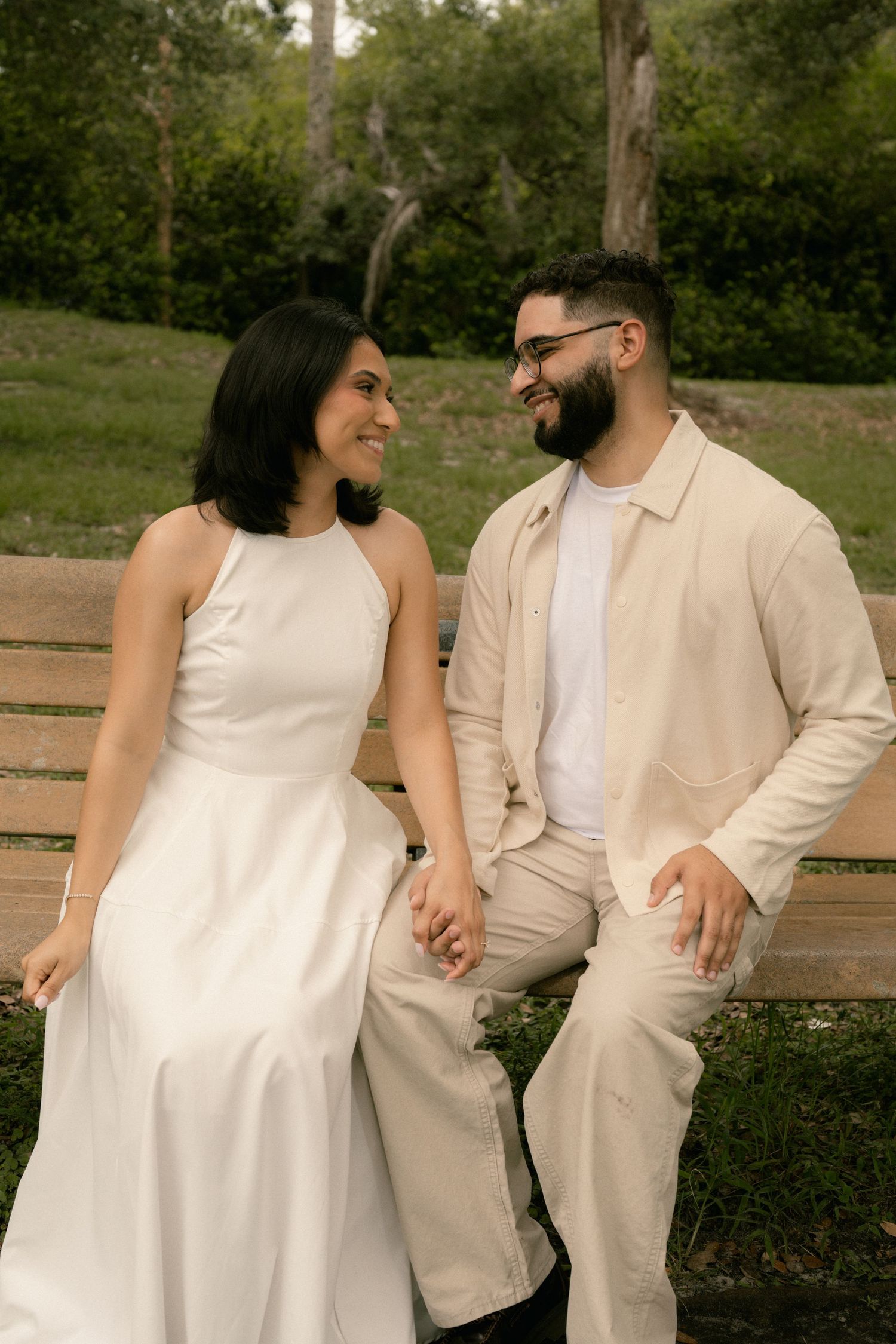 A happy couple sits on a wooden bench in a park, holding hands and smiling at each other lovingly.