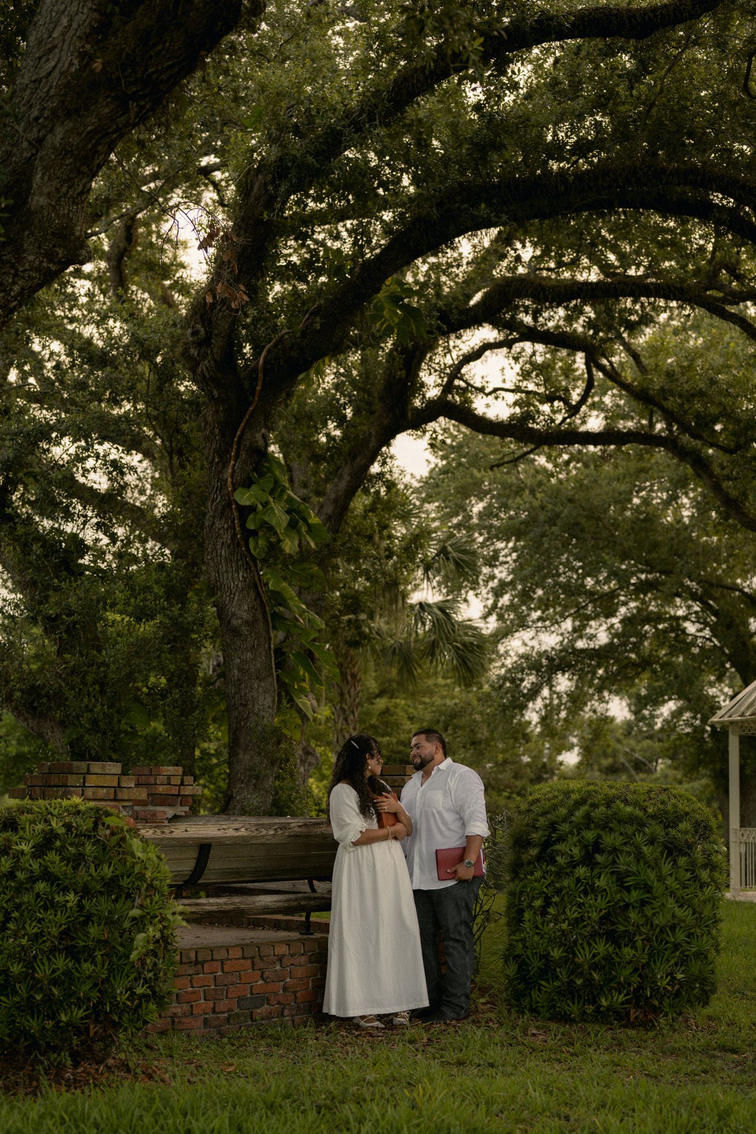Couple in white dress and shirt standing under large oak trees in a lush green park setting.