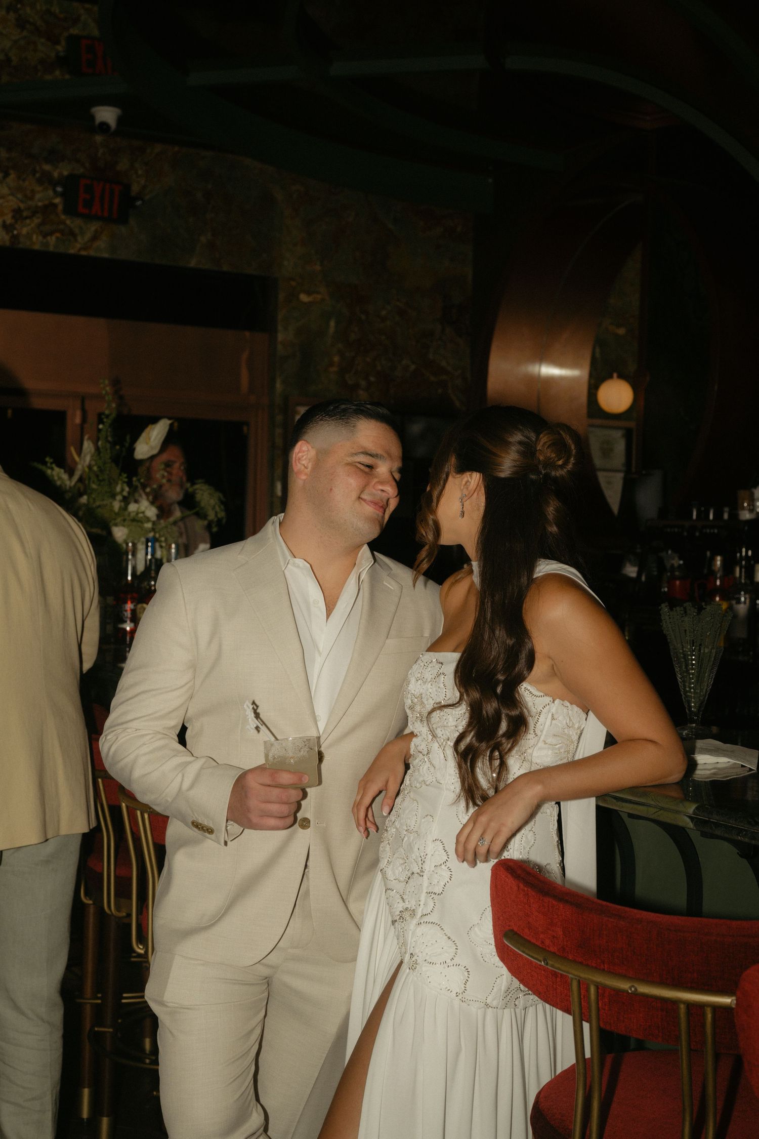 Bride and groom in white attire share a romantic moment at a dimly lit wedding reception bar.