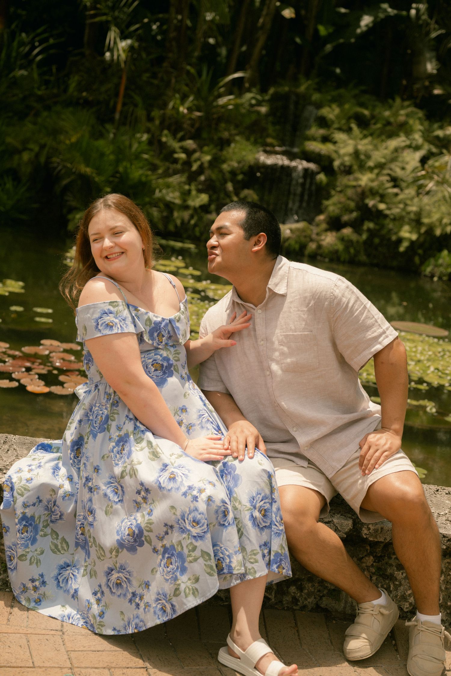 Couple sitting together by a garden pond, woman in floral blue dress, man in casual linen shirt, smiling outdoors.