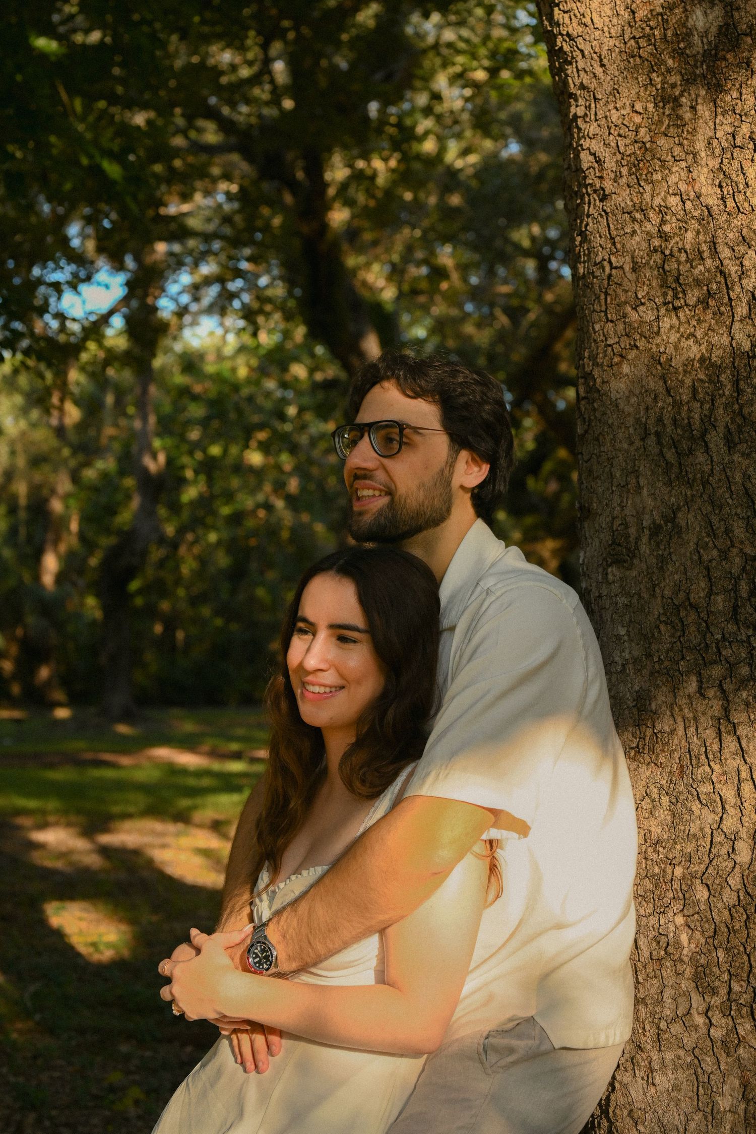 A couple embraces while sitting against a tree trunk in a sunlit park during golden hour.