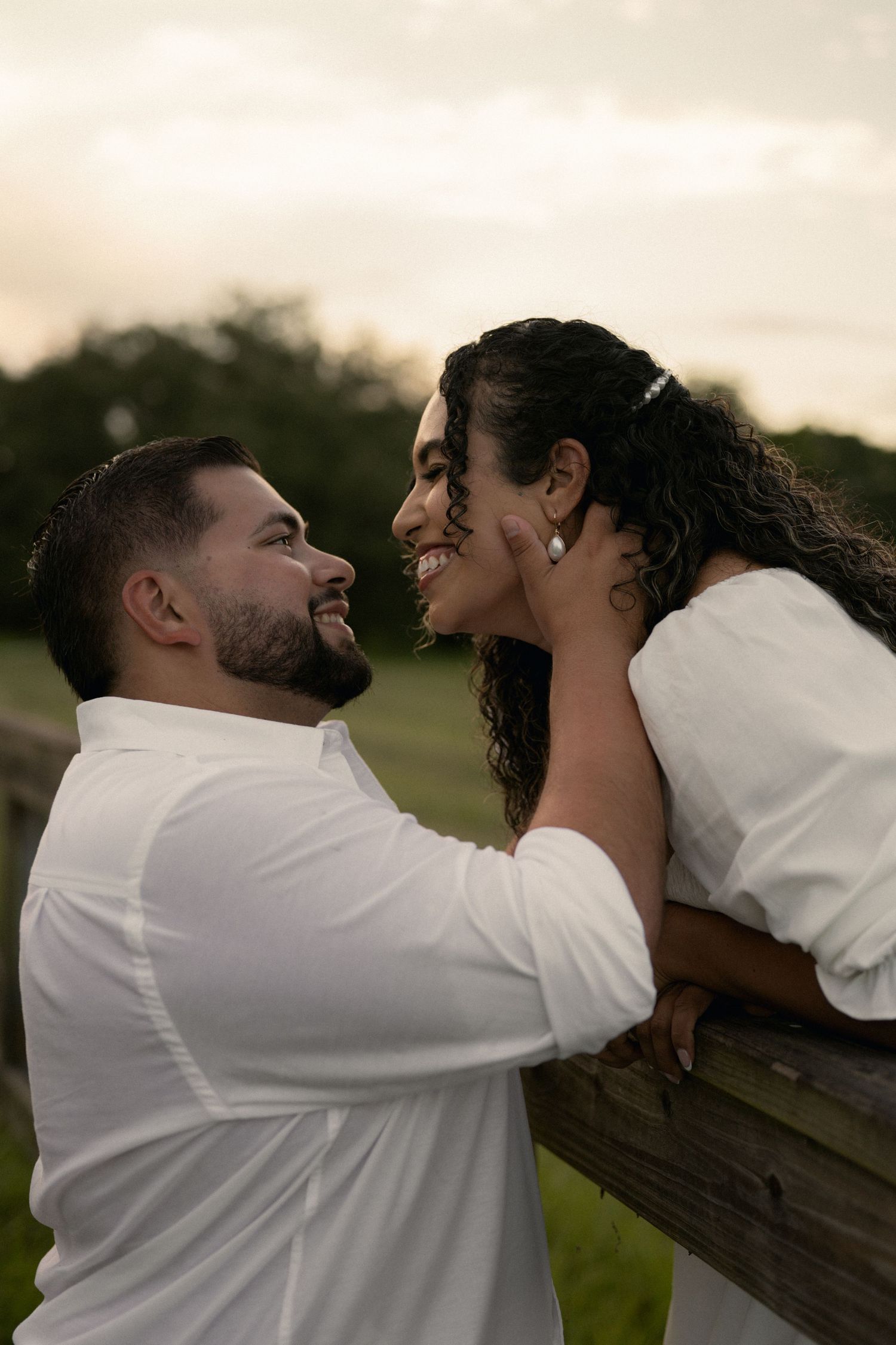 Smiling couple gazing at each other near a wooden fence at sunset, man tenderly touching womans face.