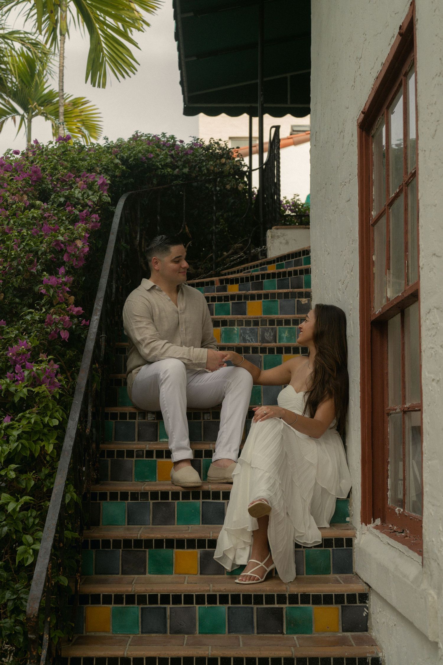 Couple in white outfits sitting on colorful tiled steps beside a building with lush tropical greenery.