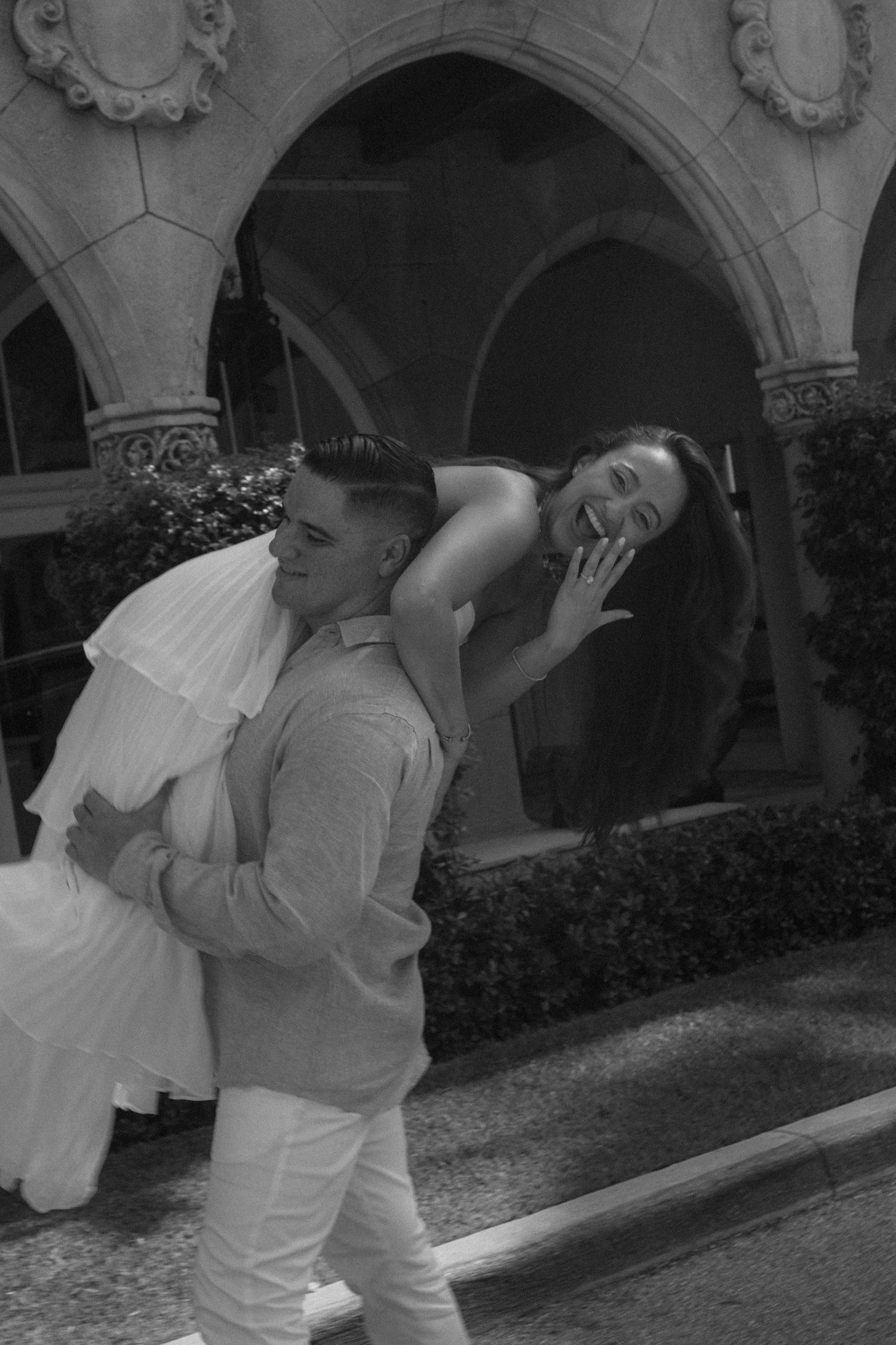 Black and white photo of a groom carrying his laughing bride on his back outside an elegant arched building.