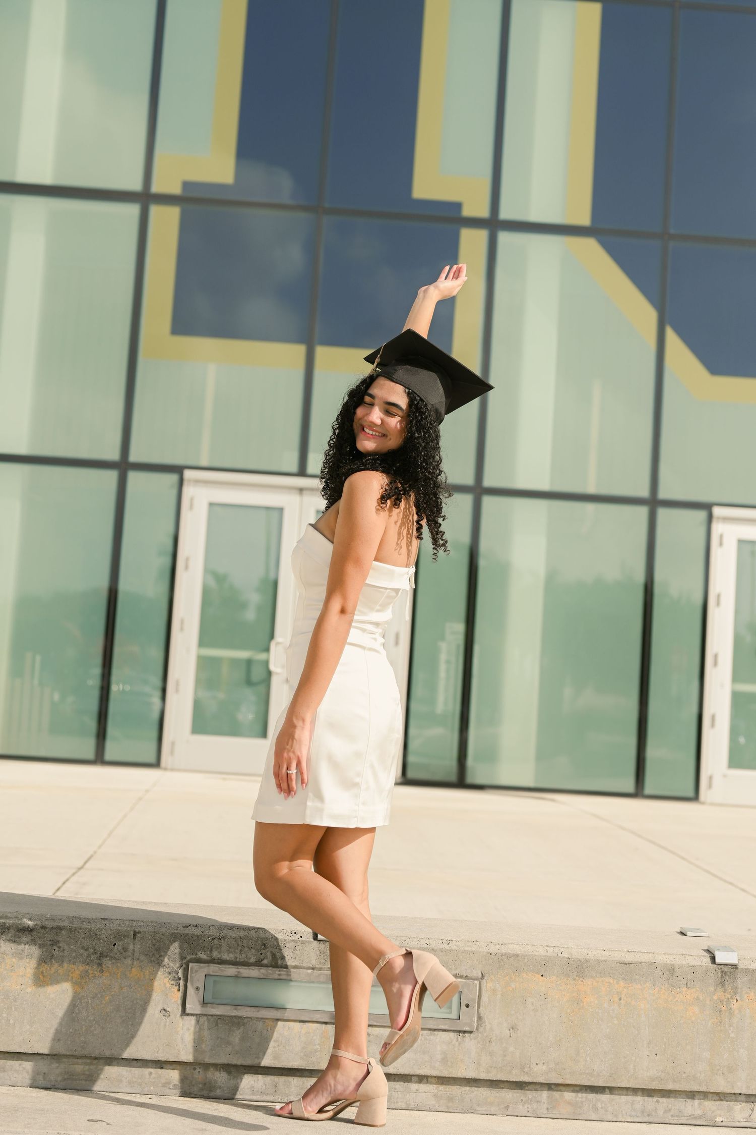 Young woman in white dress and graduation cap celebrates outdoors near modern glass building.