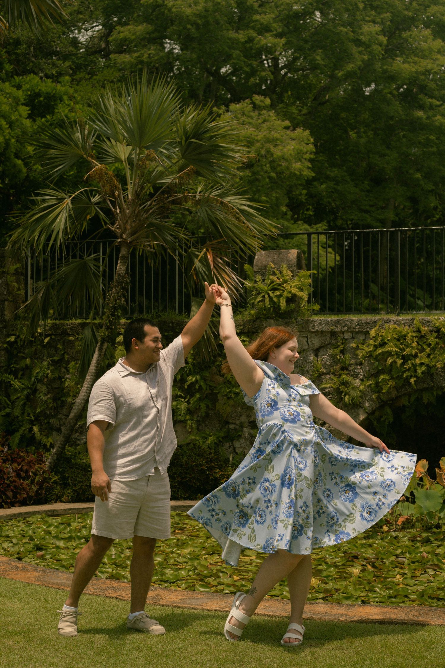 Joyful couple dancing outdoors in a tropical garden, woman twirling in a blue floral dress.