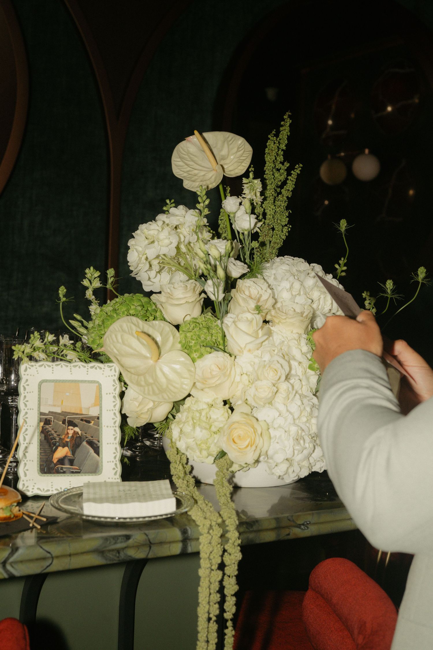 Person arranging a lush white floral arrangement with roses, hydrangeas, and anthurium on a marble table.