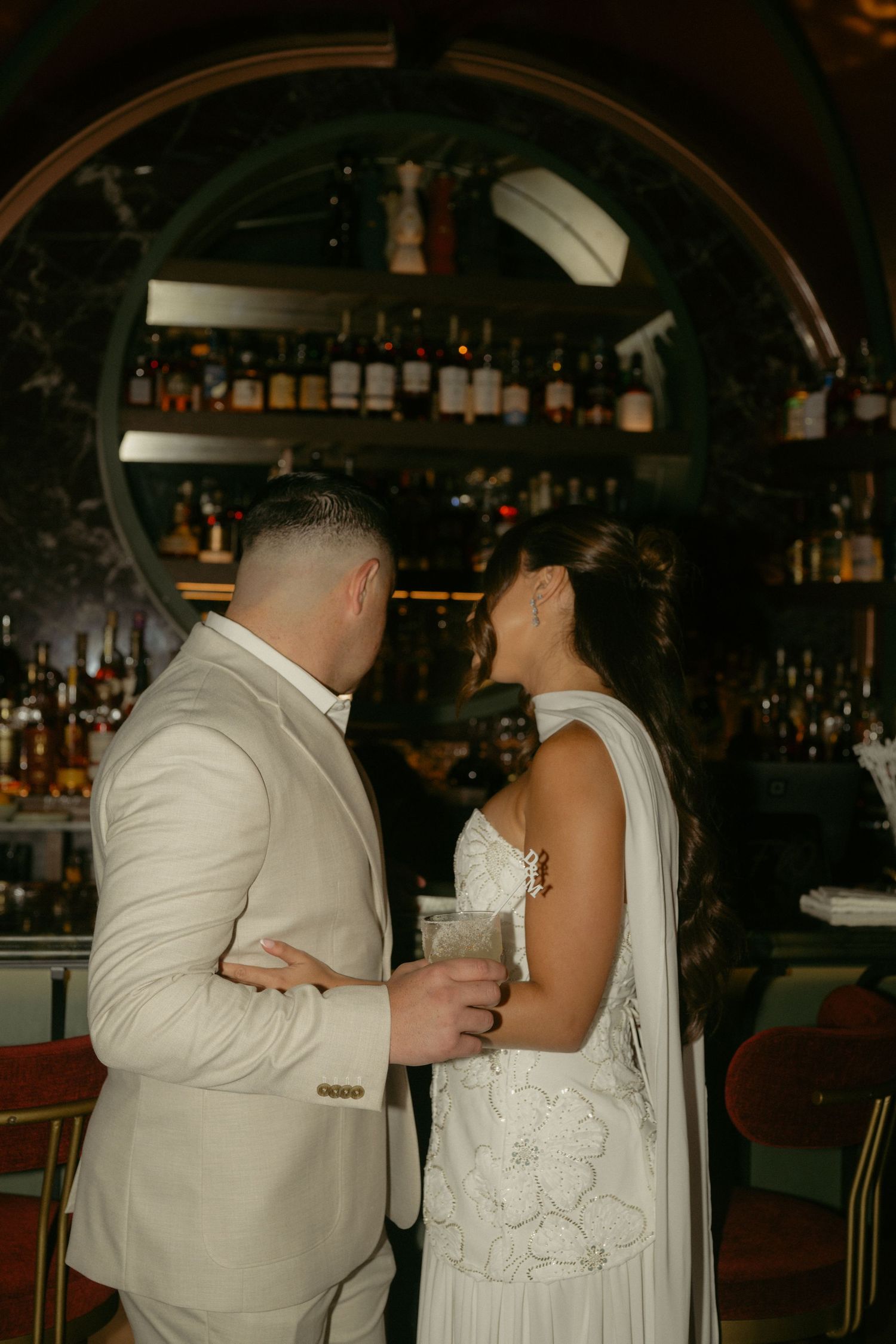 A couple in wedding attire shares an intimate moment at a dimly lit bar with a circular bottle display backdrop.