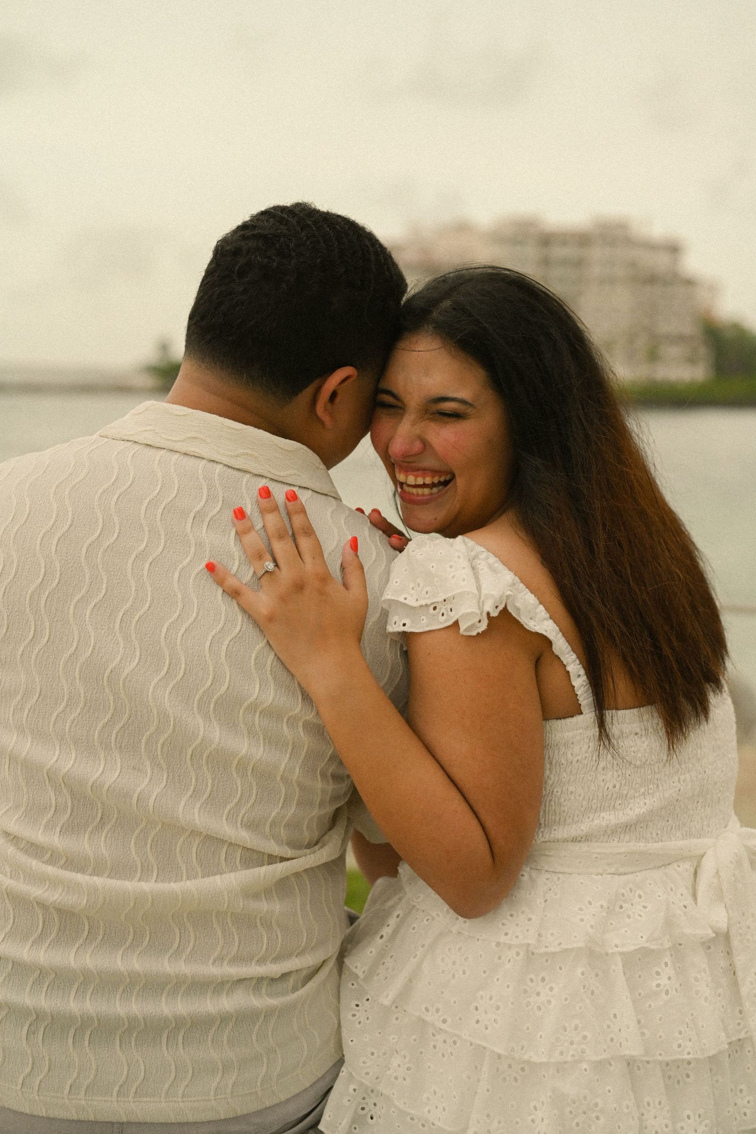 A couple in white outfits shares joyful laughter during a beachside photoshoot.