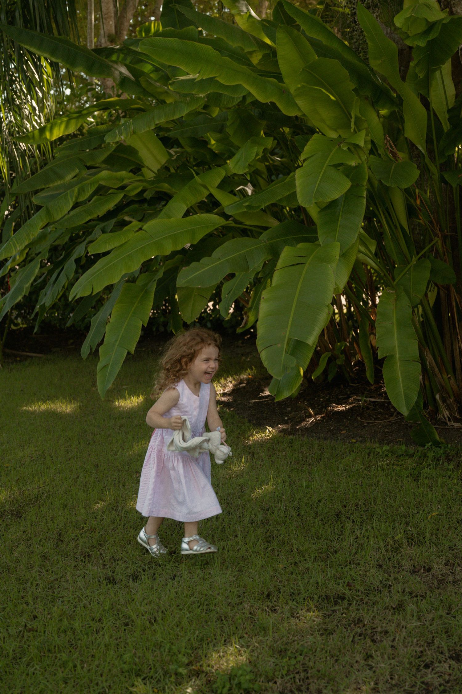 A young girl with curly hair in a light pink dress runs across a sunlit green lawn beside large tropical banana leaves.