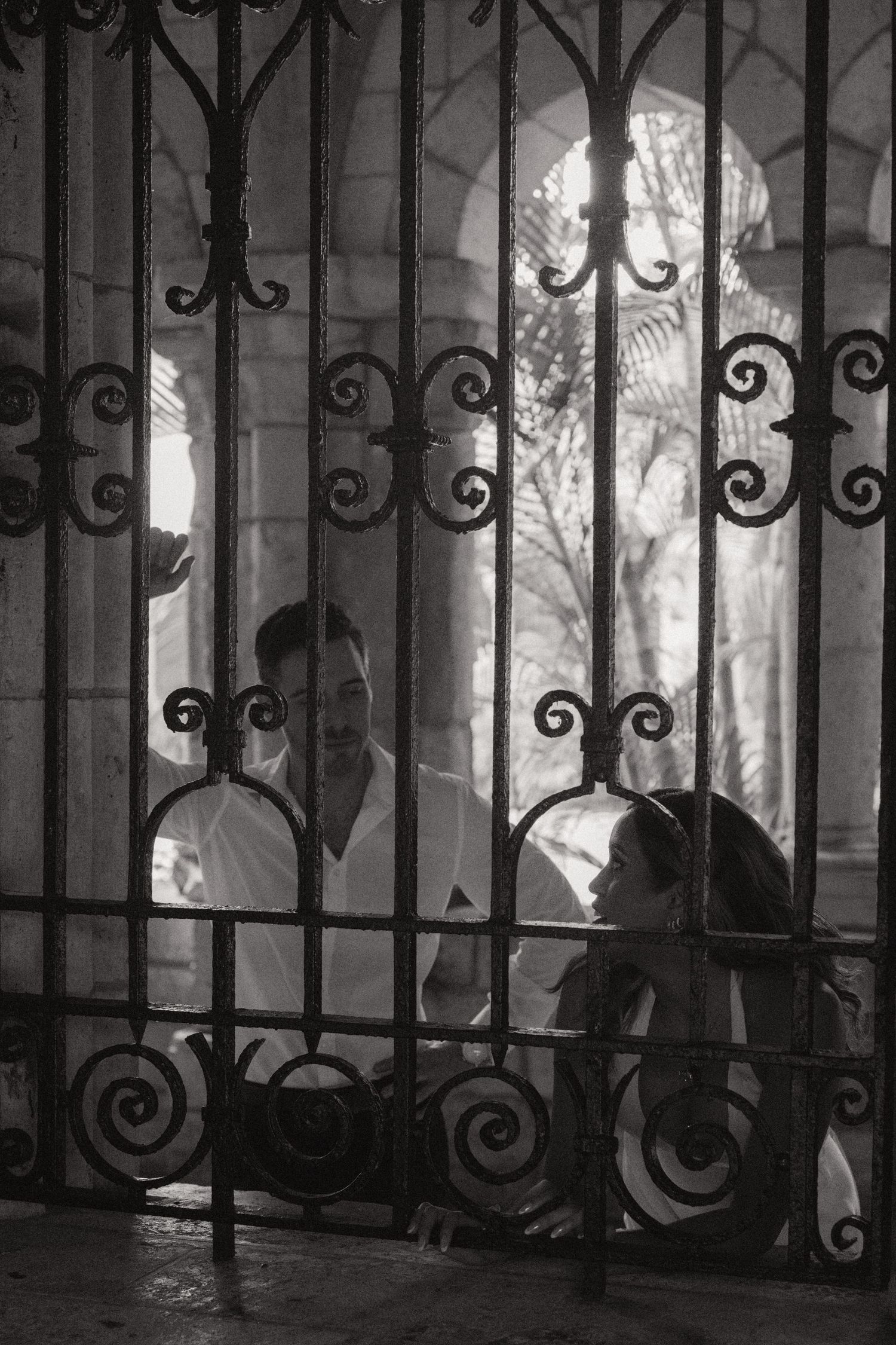 Black and white engagement photo of a couple seen through an ornate wrought iron gate with decorative scrolls and stone arches behind them.